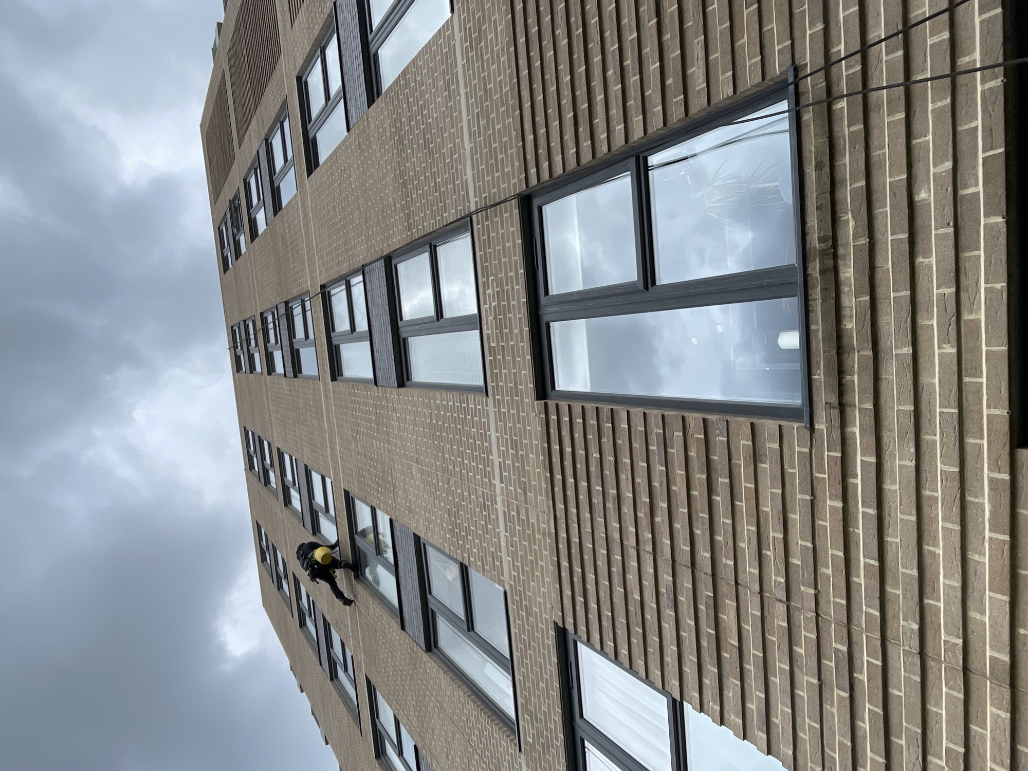 A low-angle, eye-level perspective photograph shows a window cleaner rappelling down the side of a building with brick walls and black-framed windows on a cloudy day in London, United Kingdom. The building has alternating bands of vertically-laid bricks and stacked bricks. The window cleaner, dressed in dark clothing, appears to be cleaning one of the windows, and a yellow bucket is visible near them. Dark, overcast skies are visible behind the building.