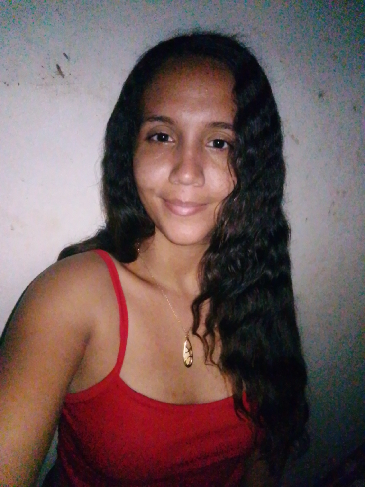 A medium shot shows a person with long, dark, wavy hair and brown skin. They are wearing a red, thin-strapped top and a gold pendant necklace. The background is a plain, light-colored wall, slightly out of focus. There is no indication of the city of Juan Griego, Venezuela.