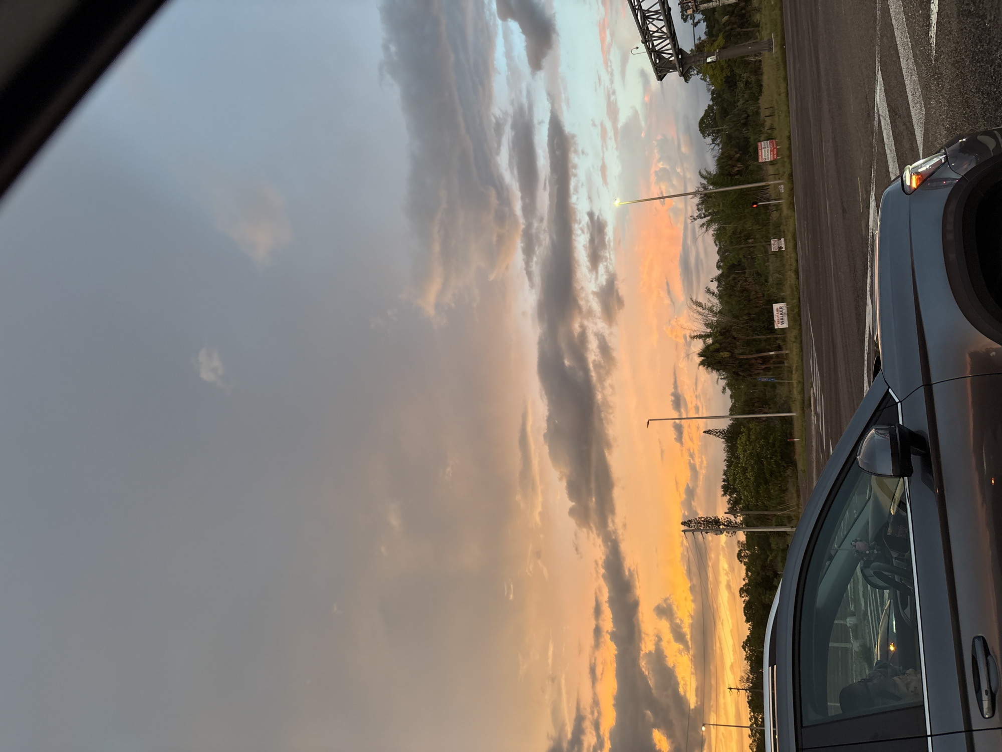 An overcast sky with vibrant sunset colors stretches over a landscape dominated by lush green trees and a road with white markings. In the foreground, the side of a dark gray SUV is visible, reflecting the ambient light. Several streetlights and signs dot the scene, with a large metal structure, possibly a bridge or overpass, in the upper right corner. The image captures a moment in what appears to be a suburban or highway environment.
