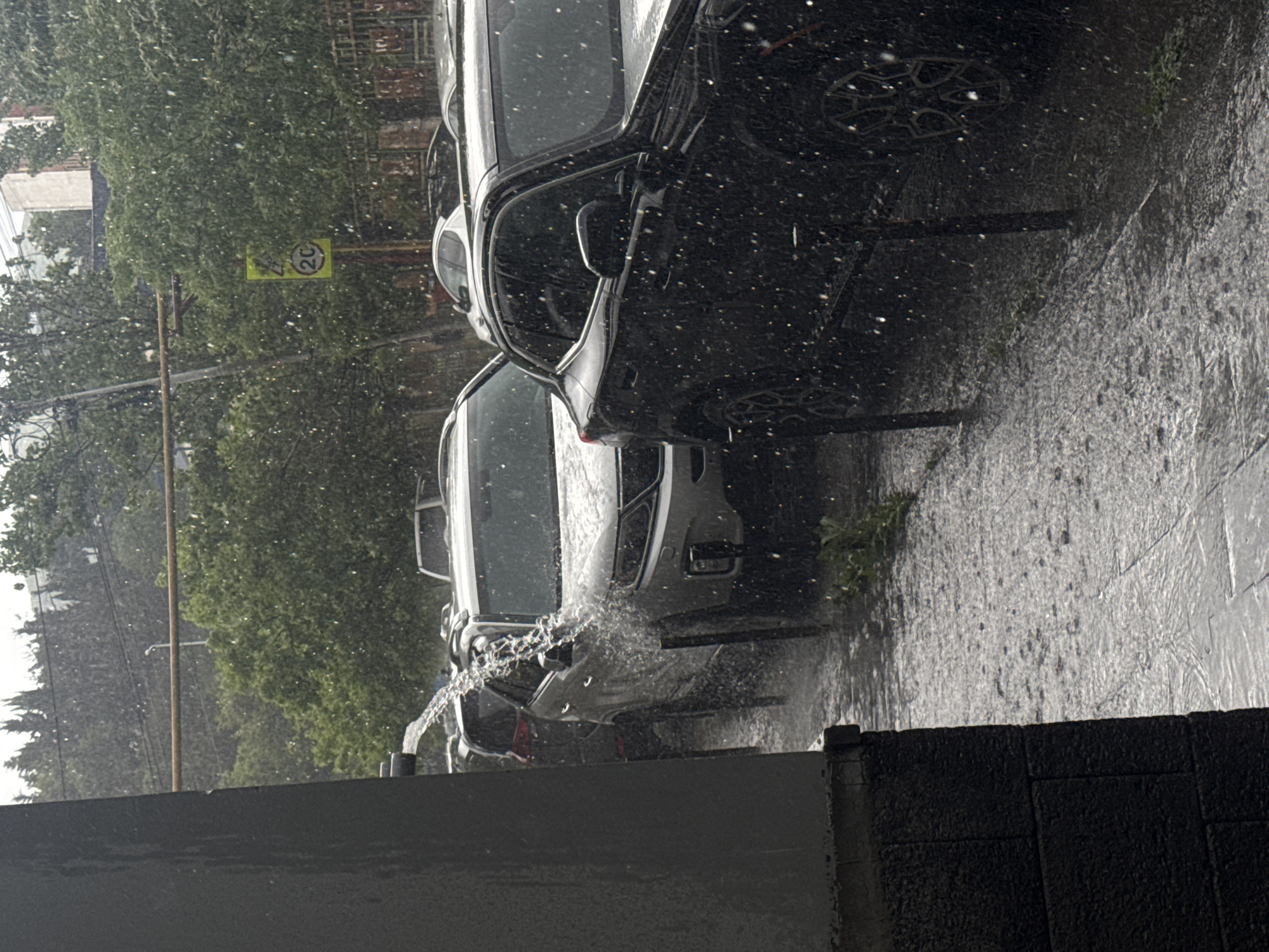 It is raining heavily in Tbilisi, Georgia. The view is from an elevated position, likely an apartment or building. Several cars are parked along the street, visible through the downpour. A speed limit sign indicating "20" is present in the background, partially obscured by foliage. Water is sheeting off the vehicles and sidewalk. The foreground shows a dark, textured wall or ledge, implying the viewpoint is inside or partially sheltered.