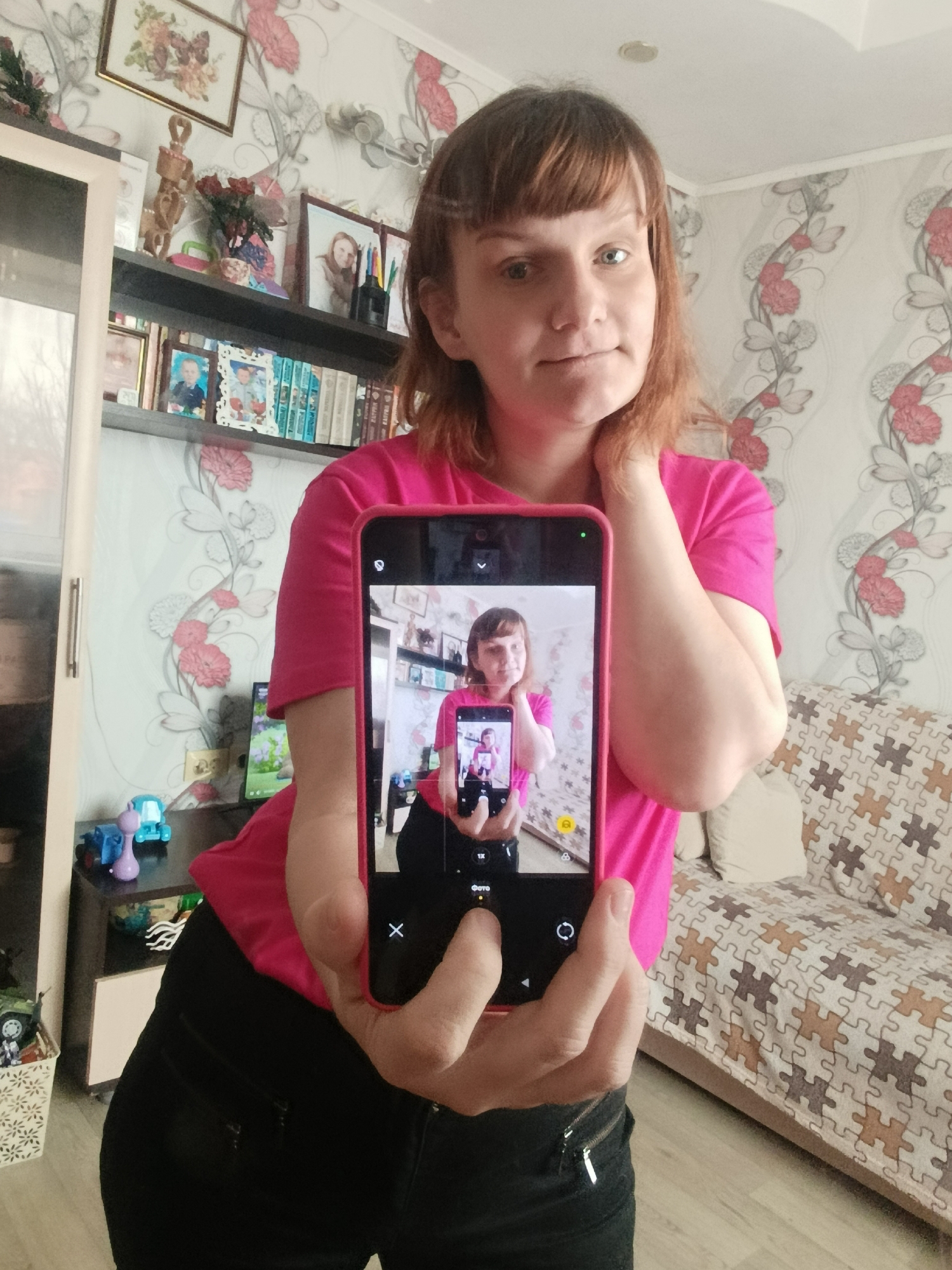A woman with fair skin and reddish-brown hair is holding a smartphone in front of herself. The phone's screen displays an image of the woman holding a smartphone, creating a "photo in photo" effect. She is wearing a bright pink t-shirt and black pants. Her right hand is raised to her neck. Behind her is a room with floral wallpaper. A shelf is visible with decorative items, including picture frames and books. A sofa with a jigsaw puzzle-patterned cover is also in the background. The location is indoors, presumably a private residence.