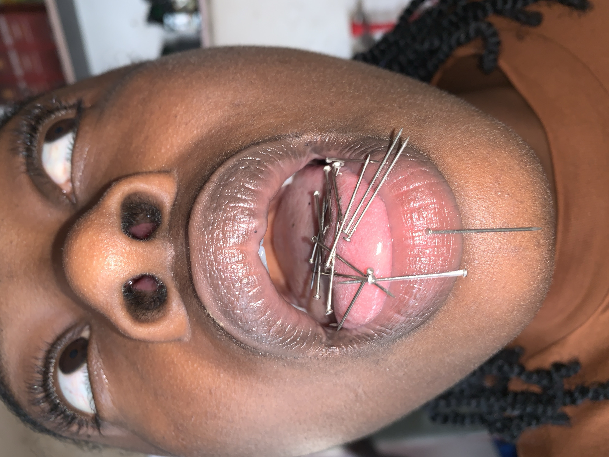 A close-up, low-angle photograph depicts a person with dark brown skin, gazing upwards. The eyes feature long, dark eyelashes and visible sclera with minor vascularization, positioned above dark irises. Dark hairs are visible within the nostrils. The mouth is wide open, revealing full, glossy lips, a pink tongue, and the interior of the oral cavity. Numerous metallic, silver-colored pins, consistent with sewing pins, are present. One pin is visibly inserted through the lingual frenulum. Multiple pins are either embedded in the tongue's surface or resting upon it, arranged in a seemingly haphazard manner. Several additional pins protrude from the lower lip, extending downwards. Dark braided hair is visible on the left side of the frame, and the person wears a rust-colored garment. The background is blurred, indicating an indoor setting with indistinct objects. This scene is located in New Stadium, Nigeria.