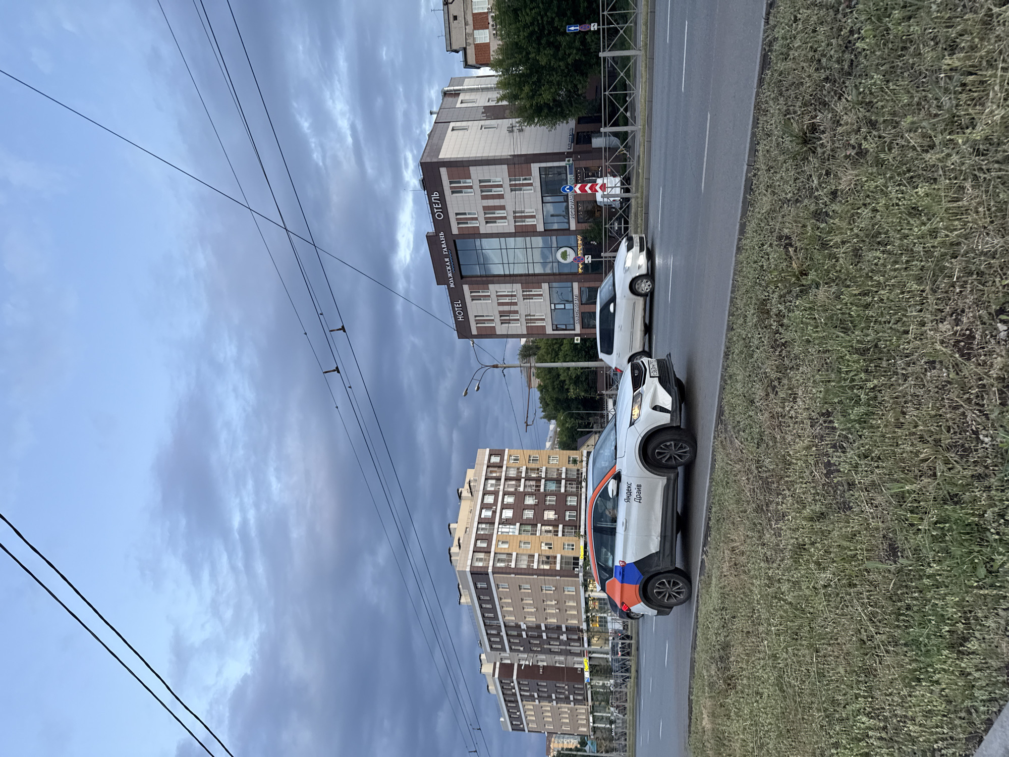 The image captures a street scene in Kazan, Russia, likely during the early evening under a cloudy sky. In the foreground, a white Yandex Drive car with orange and blue accents is driving on a multi-lane road. The car has "Яндекс Драйв" text visible on its side. Further down the road, a white hatchback is also in motion.
On the right side of the road, a modern hotel building with the text "HOTEL ВОЛЖСКАЯ ГАВАНЬ" (Hotel Volga Haven) is prominently displayed. The building features large glass windows and a facade with brown and beige panels. Various signs, including one for "РЕСТОРАН" (Restaurant), are visible on the lower level. Adjacent to the hotel, a portion of another building can be seen.
On the left side of the street, two multi-story apartment buildings with a similar architectural style, featuring balconies and many windows, are visible. A row of trees lines the area between the road and the buildings. Overhead, power lines crisscross the sky, which is filled with dramatic, dark clouds. The scene appears to be a typical urban environment with traffic and residential and commercial buildings.