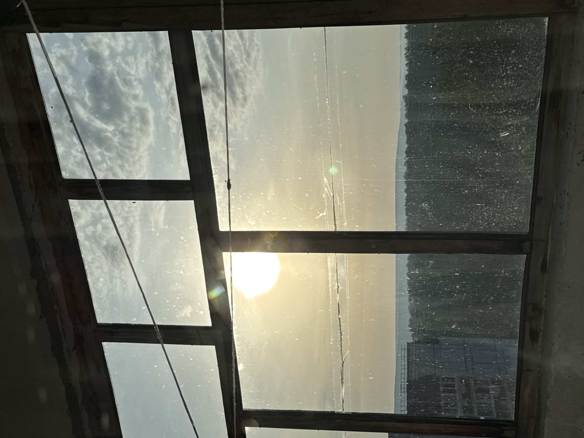 The image is a low-angle view through a multi-pane window in a likely indoor setting, possibly a loft or attic space. The wooden window frame divides the view into several rectangular sections. The sunlight is shining directly through the window. The sky is partly visible through the panes with some cloud cover. In the background through one of the panes, a building is reflected. The pane with the bright light shows some cracking and wear. The background appears slightly hazy. The location is Chaikovsky, Russia.
