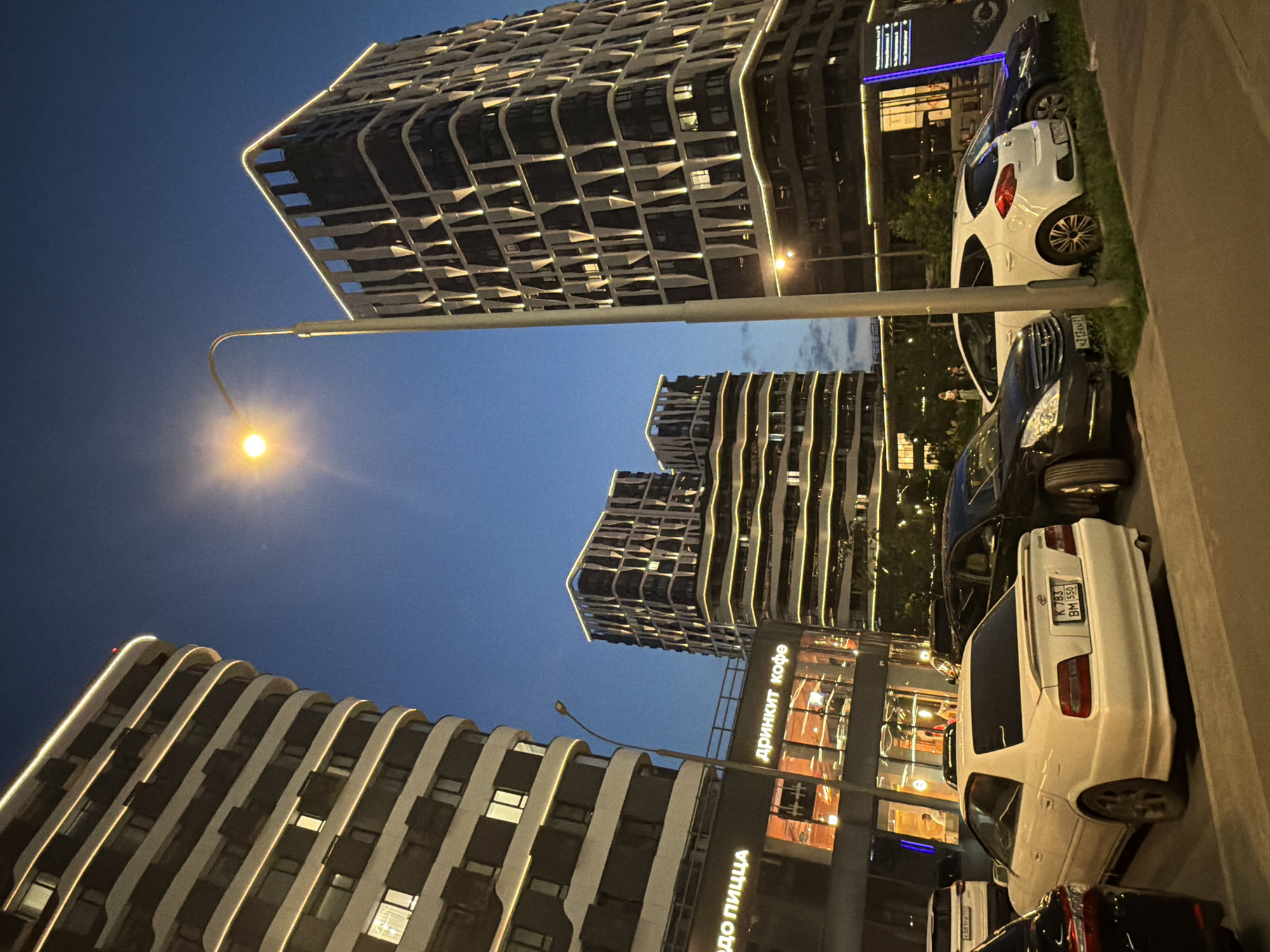 The image depicts a street scene at dusk in Moscow, Russia, specifically in the vicinity of the Moscow City business district. Two tall, modern buildings with illuminated outlines are prominently featured. One building on the left has a curved facade and is lit with a continuous, thin yellow light along its horizontal edges. The building on the right has a more angular design, with vertical decorative elements and similar illuminated horizontal trim.
In the foreground, several cars are parked. The most visible car is a white sedan, possibly a Toyota Mark II, with a license plate that appears to be from Russia. Other cars visible include a dark sedan and a white hatchback. A street lamp with a single glowing light stands between the two buildings, casting a warm illumination.
Below the left building, there are storefronts with illuminated signs. One sign clearly reads "Дринкит кофе" (Drinkit Coffee), and another partially visible sign says "ДО ПИЦЦА" (Do Pizza). The overall atmosphere is that of a modern urban environment at the transition from day to night, with artificial lighting playing a significant role. The sky is a deep blue, indicating late evening or early morning.
