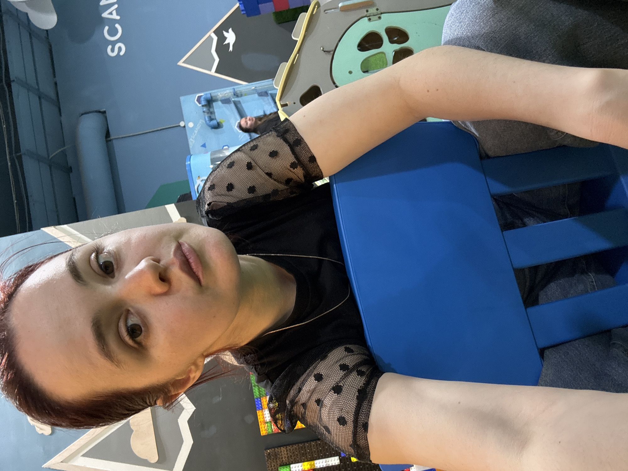 An angled selfie shows a person, likely a woman, in an indoor setting resembling an interactive play area or children's museum in Izhevsk, Russia. The subject is in the foreground, looking directly at the camera with a neutral expression. She has auburn hair, dark eyes, and light skin, and is wearing a black top with sheer, polka-dotted sleeves and a thin chain necklace. Her arm is propped on a bright blue chair or piece of furniture. Dark trousers or jeans are also visible on her lower body.
The background features a blue wall with large white letters spelling "SCAN" partially visible. Below this, a light blue pipe-like structure is present. Further back, another person with dark hair is lying down on a blue surface. To the right, a large beige and light green play structure with various cutouts is seen. At the bottom of the frame, a colorful mosaic made of interlocking blocks, possibly LEGOs, is visible. Another white geometric design is near the subject's head. The indoor lighting suggests it is daytime. Faint text, "SCANSYS", can also be discerned on a blue object in the mid-ground. The overall scene suggests a vibrant, engaging environment.