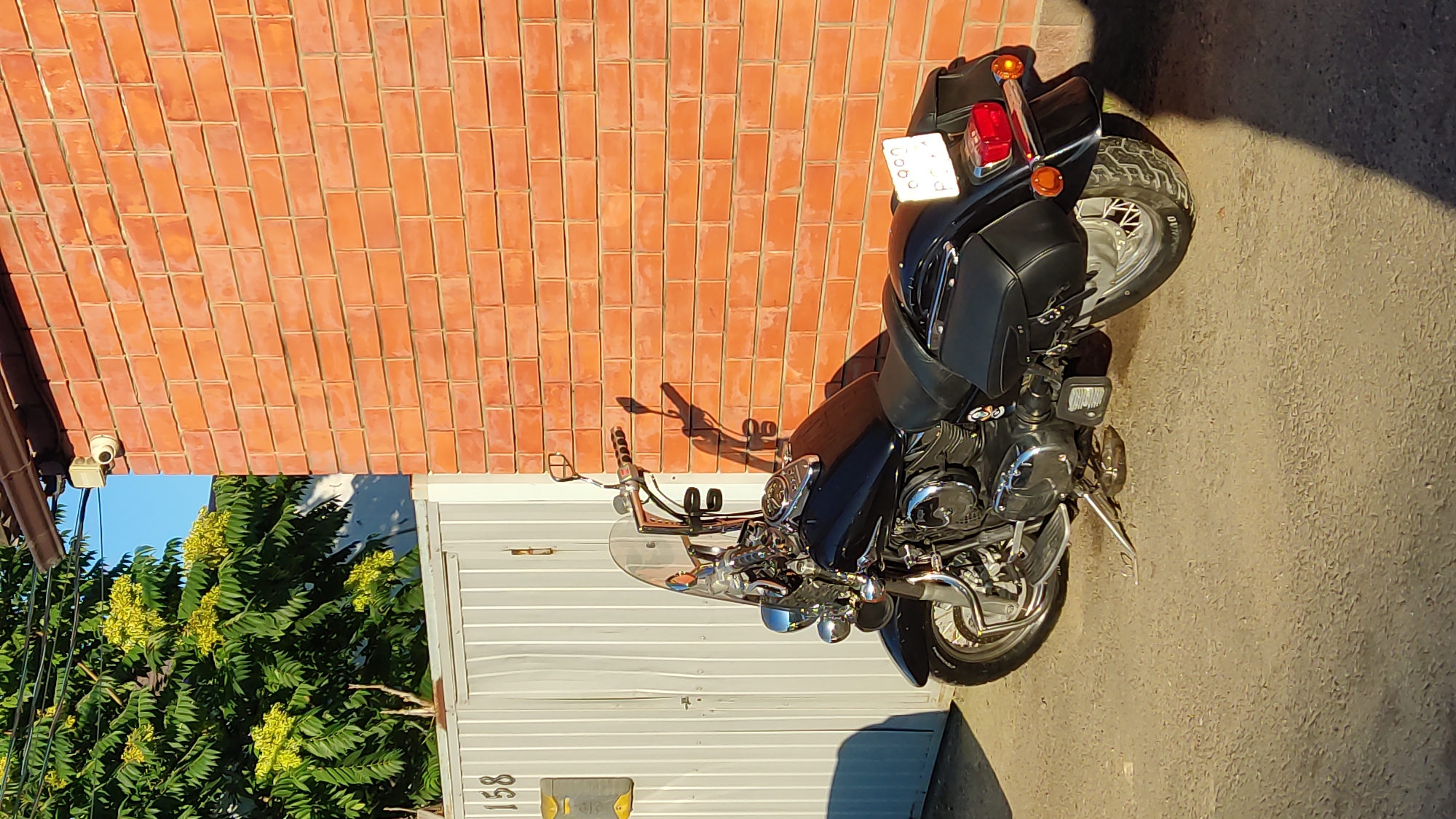 A black motorcycle is parked on a paved surface next to a red brick wall. The motorcycle has a windshield, saddlebags, and chrome accents. A white gate is visible behind the motorcycle, with the number "158" on it. A security camera is mounted on the brick wall. The scene appears to be outdoors, with sunlight casting shadows. The image was taken in Krasnodar, Russia.