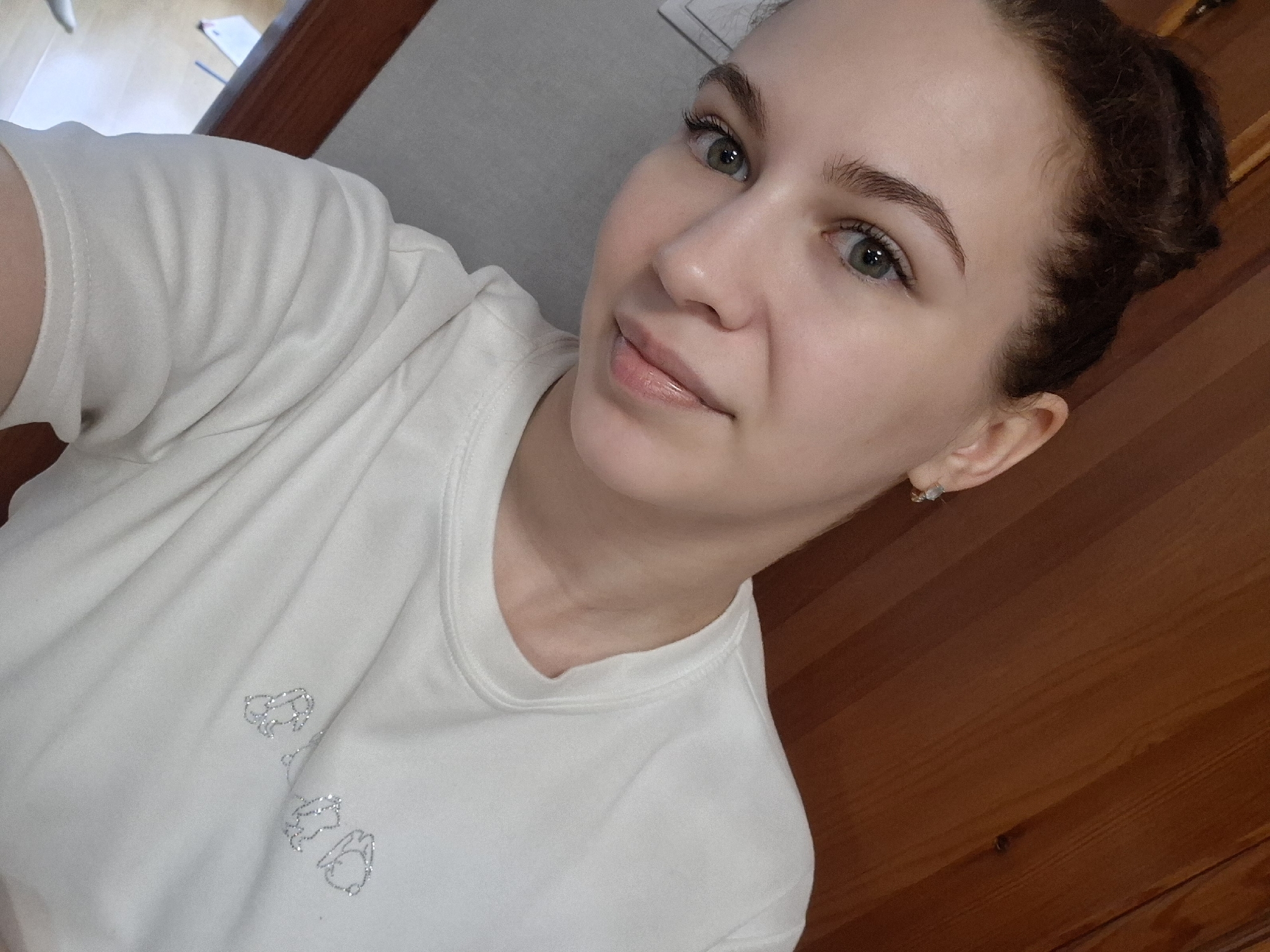 A woman with light skin, green eyes, and brown hair pulled back into a bun is taking a selfie indoors. She's wearing a white t-shirt with a simple design near the neckline. Behind her is a wooden wall and a light-colored wall. The location is likely in Chaikovsky, Russia.