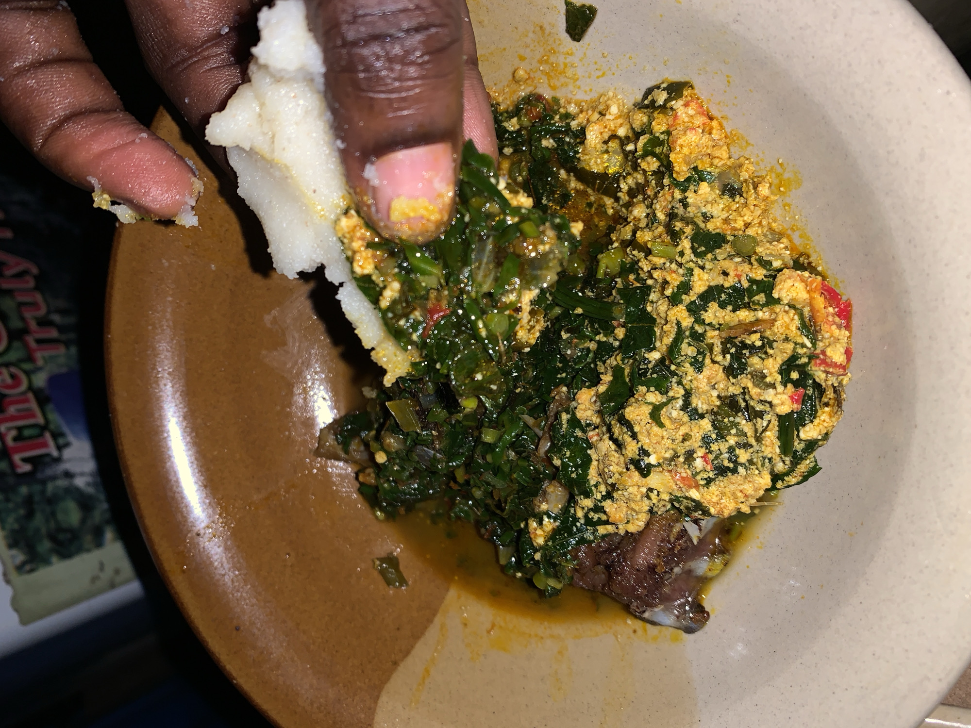 A close-up shot depicts a hand holding a piece of a white, doughy food, presumably a staple like fufu or pounded yam, and dipping it into a bowl of stew. The stew appears to be a mix of green leafy vegetables, a yellow, scrambled-egg-like substance, and reddish pieces that could be tomatoes or peppers. There seems to be a piece of fish at the bottom of the bowl with brown skin. There is also some visible liquid or oil in the bowl. The food is served in a wide, shallow bowl, with a dark brown edge. The exact location in the city of Kabong, Nigeria is unknown based on the image.