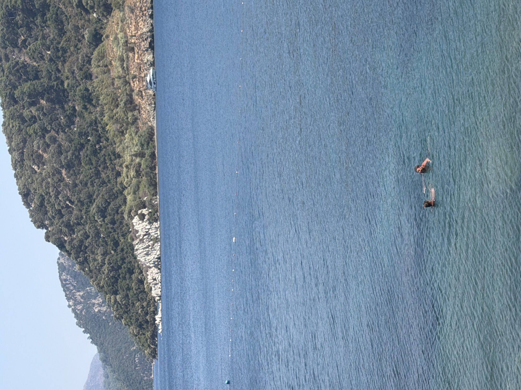 Two individuals are swimming in the clear blue waters of Marmaris, Turkey. The sea is calm, with gentle ripples reflecting the sunlight. In the distance, a lush, tree-covered hillside slopes down towards the water, punctuated by rocky outcrops. A white yacht is moored near the shore, adding a touch of luxury to the natural landscape. A string of buoys marks a designated swimming area in the middle ground. The sky is a clear, bright blue, indicating a sunny day.