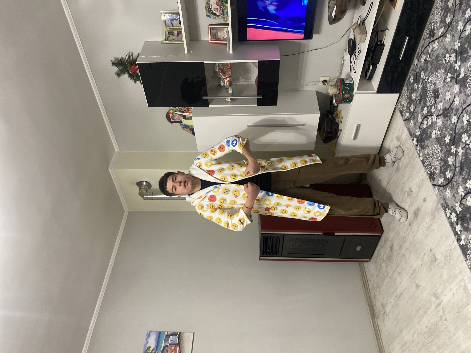 A man stands in an indoor setting, wearing a white hoodie covered with emoji graphics. He is positioned between a red and black cabinet-like structure and a white TV stand with black accents. On the TV stand, a television is visible, showing an indistinct image. The background features a white wall and ceiling, along with decorative items on the TV stand and shelves. A framed picture hangs lower on the wall. Based on the location details provided, this scene is set in Goritsa, Bulgaria.