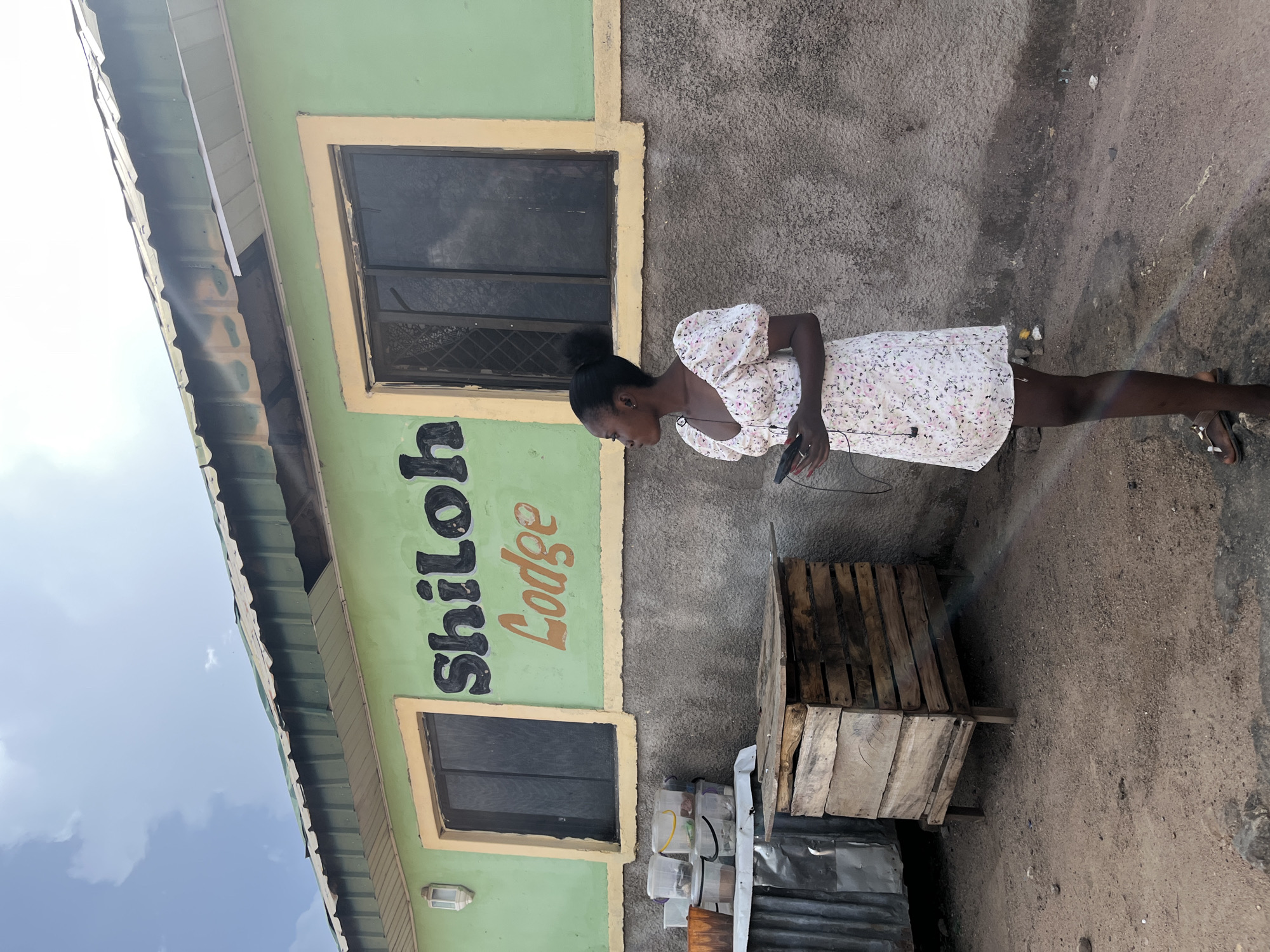 In this full-shot outdoor image, a young Black woman stands on a dirt ground in front of a light green building with a faded yellow trim around its windows. The building has a metal roof. Large black lettering on the building reads "Shiloh Lodge" in an orange script.
The woman, with her hair pulled back in a bun, wears a white dress with a floral pattern of pink and purple flowers. She is looking down and to her right, holding a black cell phone in her right hand. She is also wearing sandals. To the right of the woman are two wooden crates, one on top of the other. To the left of the woman, stacked plastic containers sit on a metal shelf.
The scene suggests a daytime setting, with bright sky visible in the upper left of the image. The overall impression is of a somewhat weathered, possibly commercial building.