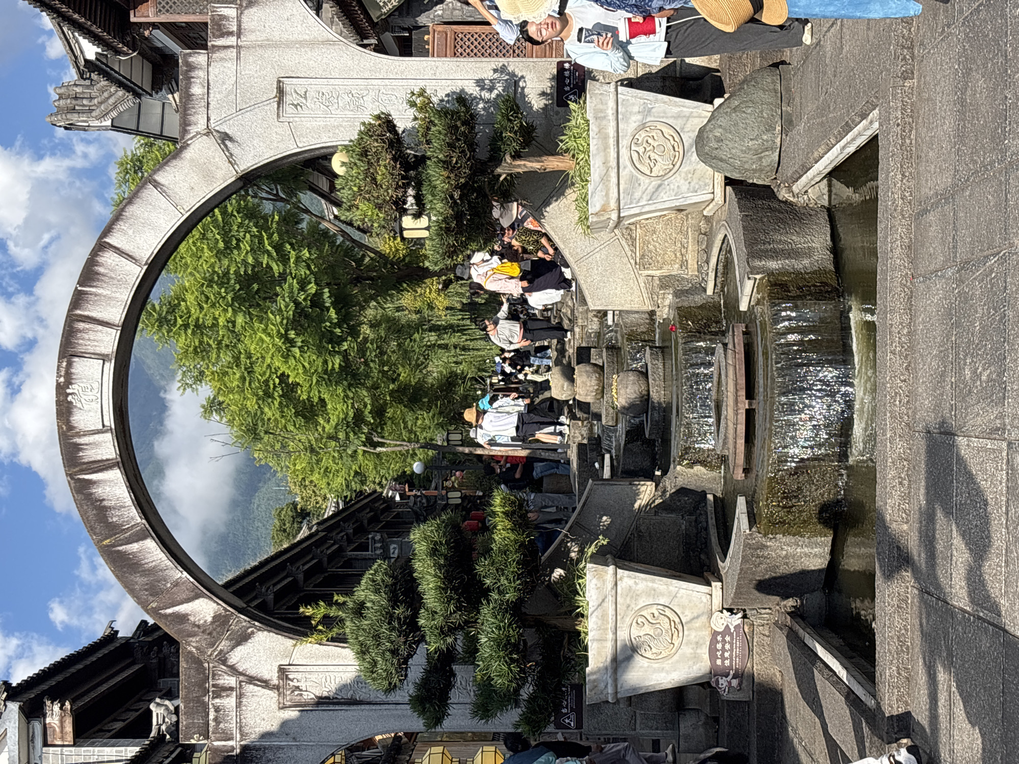 The image captures a scene in Dali City, China, featuring a traditional stone archway and a multi-tiered water fountain. The archway, made of weathered stone, frames a view of lush green trees and distant mountains shrouded in mist. On either side of the arch, traditional Chinese architecture with tiled roofs is visible.
In the foreground, a stone fountain cascades water down several levels into a central pool. Two large, decorative planters, each containing a meticulously pruned evergreen tree, flank the fountain. These planters are adorned with carved circular emblems. People are seen walking through the area, some ascending stairs next to the fountain and others walking beneath the archway. The ground is paved with stone tiles, and shadows from the structures and people are cast across the scene. The overall atmosphere suggests a public space, possibly a historic street or a scenic spot within Dali City.