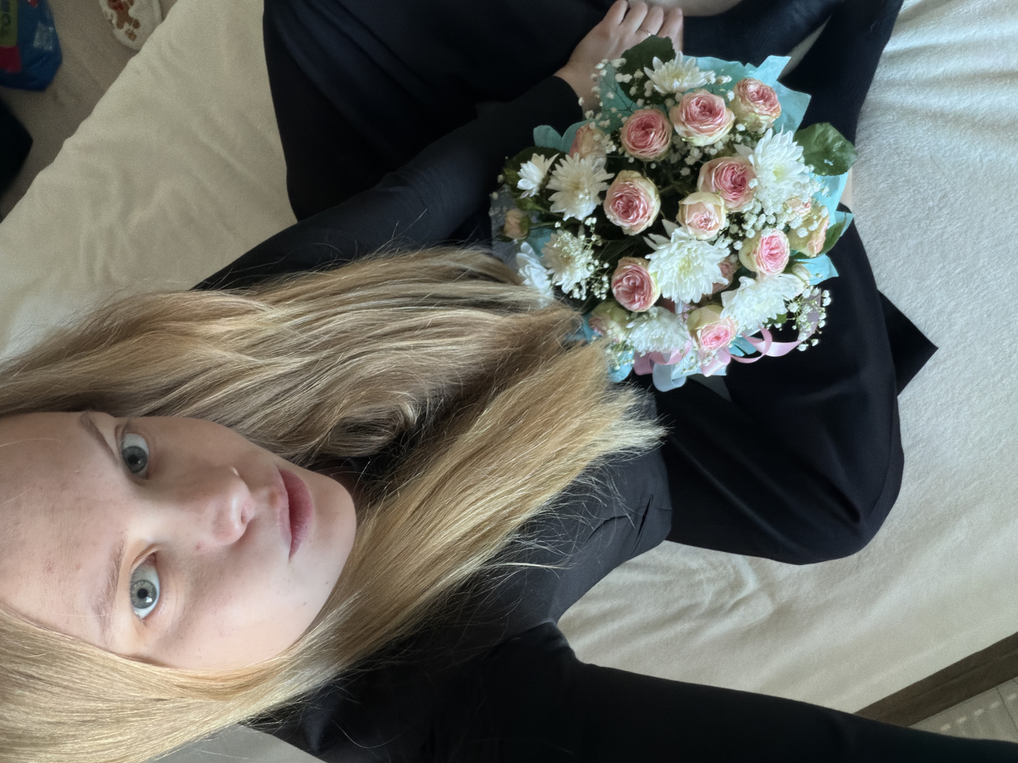A young woman with blonde hair and light blue eyes is lying on a bed, holding a bouquet of pink roses and white chrysanthemums wrapped in light blue paper. She is wearing black clothing. The location is indoors, likely a home, on a bed with white linens. There is no information in the picture that identifies the location as Izhevsk, Russia.