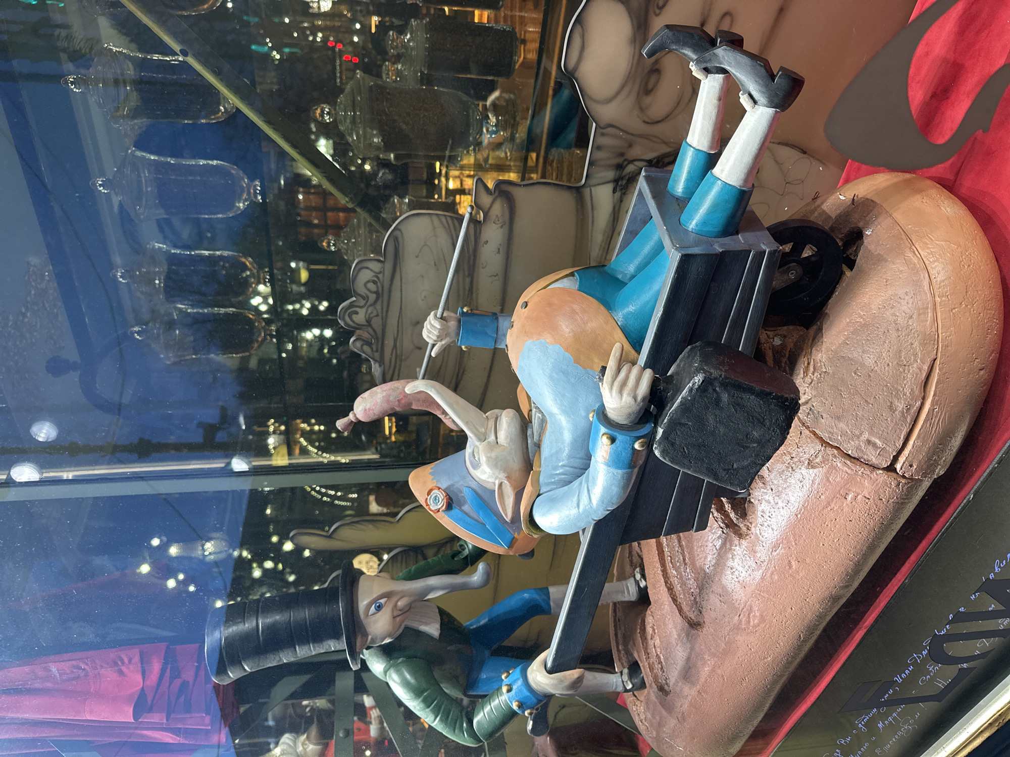 The image shows a whimsical display in a shop window, likely in Saint Petersburg, Russia based on the Russian inscription. Two figures are prominently featured: a rabbit-like character riding in a cart being pulled by a man in a top hat. The cart appears to be riding on an enlarged, vaguely anatomical object, possibly a foot or nose. Jars are visible on shelves in the background, as well as a building reflecting in the glass.
