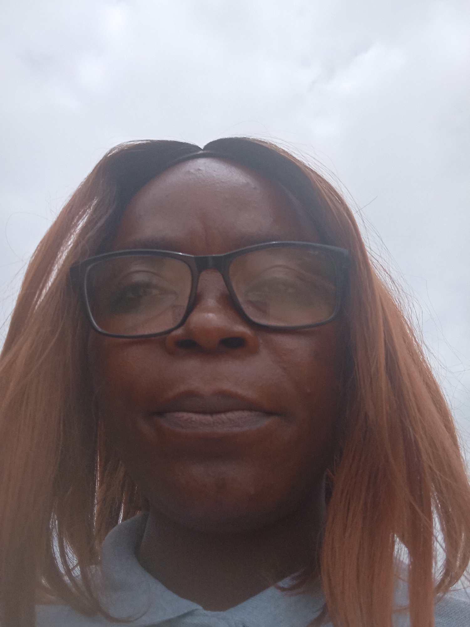 The image is a close-up, waist-up, outdoor, selfie-style photograph of a Black woman with medium-dark skin, wearing glasses and a light blue collared shirt. She has medium-length, auburn-colored hair that is parted in the middle. The background is an overcast sky. The photo was taken on a cloudy day in Kason, Nigeria.