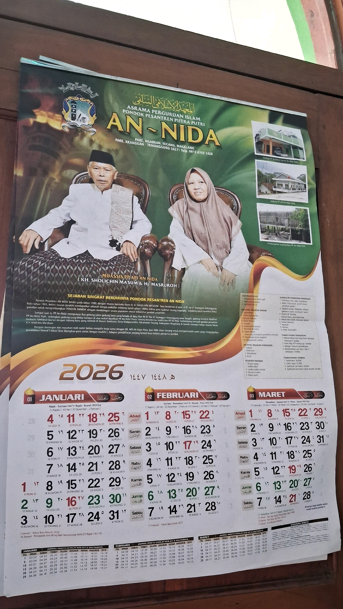 The image is a photograph of a 2026 calendar poster for Pondok Pesantren An-Nida, located in Giriwarno, Indonesia. The poster features portraits of an elderly man and woman seated side-by-side, likely the founders or prominent figures of the pesantren. The background is a lush green foliage with images of the pesantren's buildings in the upper right corner. The calendar itself is displayed prominently at the bottom of the poster, showing the months of January, February, and March 2026 with corresponding Gregorian and Hijri dates. Text in Indonesian describes the history of the pesantren and its educational programs. The overall scene is that of a promotional poster for an Islamic boarding school.