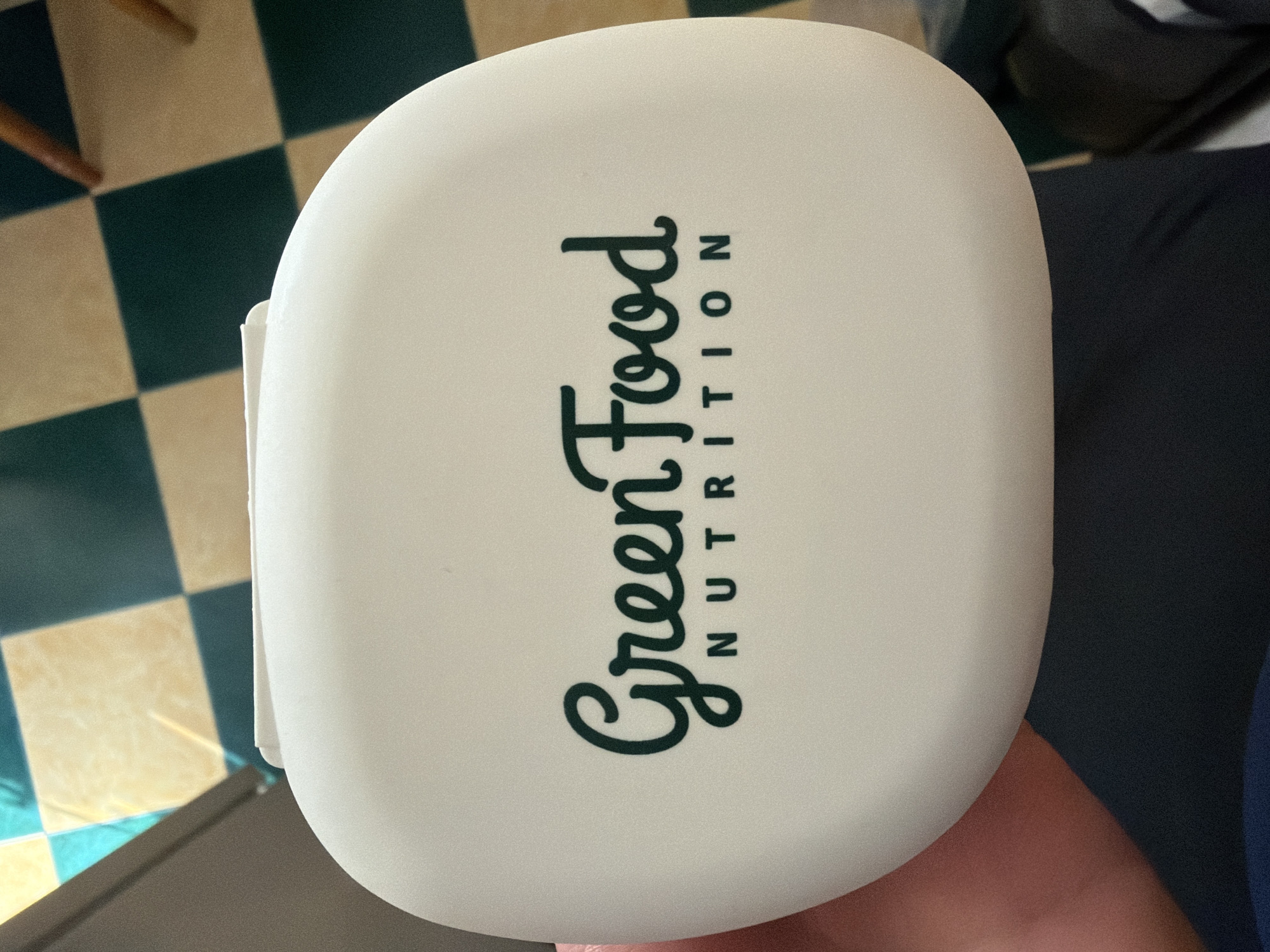 A close-up, overhead shot shows a white, oval-shaped container with the logo "Green Food NUTRITION" printed in dark green. The container is held in a person's hand, indicated by a pinkish thumb and fingers on the left side of the image. The background is a checkered floor with alternating beige and dark green tiles. Some light blue and brown elements are also visible in the background, possibly from furniture or wall decor. The lighting suggests an indoor setting.