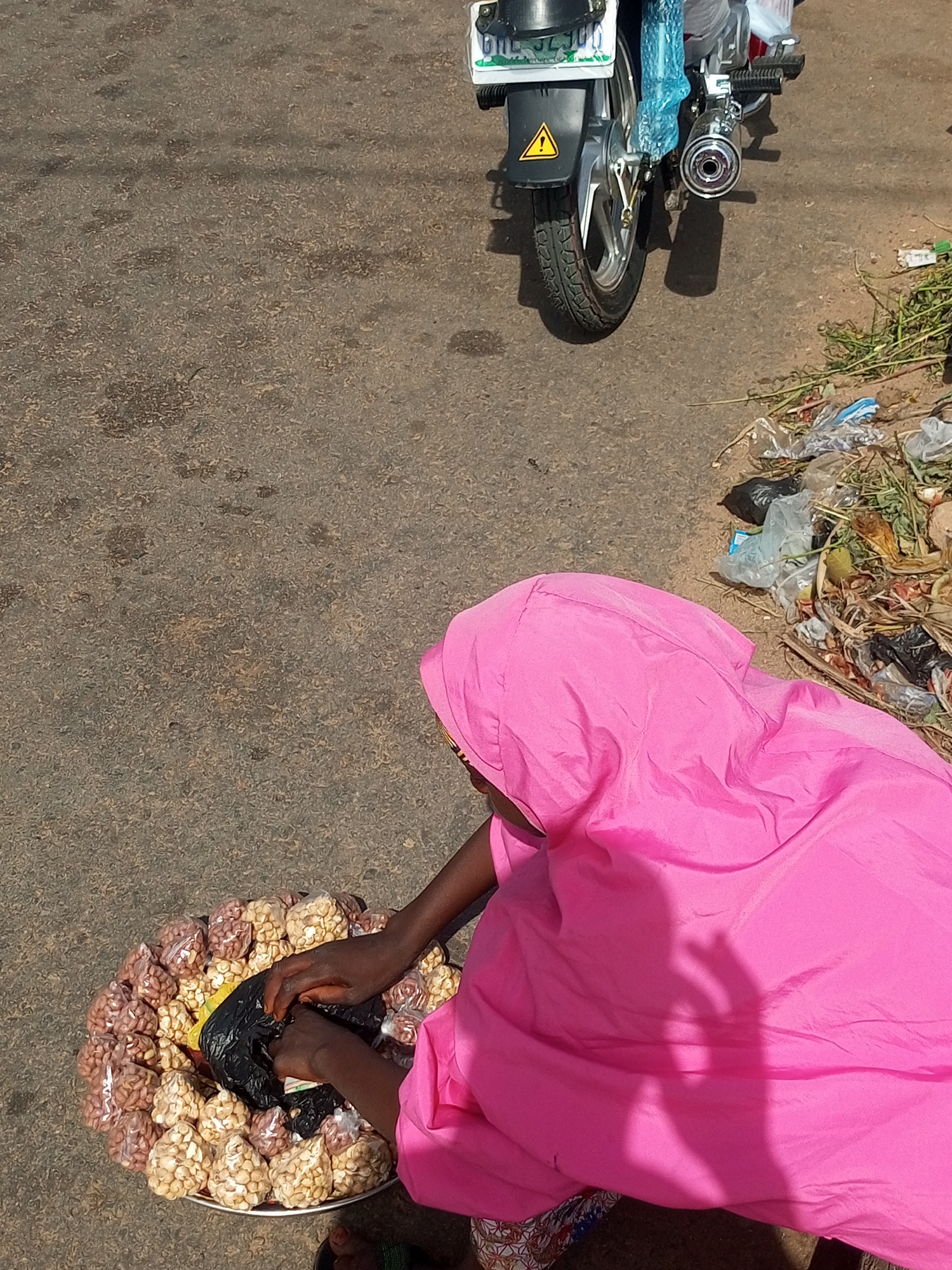An overhead shot captures a street vendor in Tempure, Nigeria, crouched next to a large circular tray filled with numerous small, clear plastic bags of peanuts. The vendor wears a bright pink hooded garment, and their dark hands are visible, seemingly arranging a black plastic bag amidst the packaged peanuts. Some peanuts appear roasted and light-colored, while others are reddish.
In the upper background, a silver and black motorcycle is partially visible, parked on a textured, grey asphalt or dirt road. Parts of the motorcycle, like the seat and some chrome, are still covered in blue protective plastic, suggesting it might be new. A license plate, partially obscured but showing "006-JL 300", is affixed to the rear fender, which also features a yellow warning triangle sticker. The chrome exhaust pipe is prominently displayed.
To the right of the motorcycle, a messy pile of roadside trash, consisting of plastic bags, dry leaves, and other organic matter, is accumulated. The scene is brightly lit by strong sunlight, casting distinct shadows from the vendor and the motorcycle, indicating it is daytime. The ground appears rough and uneven.