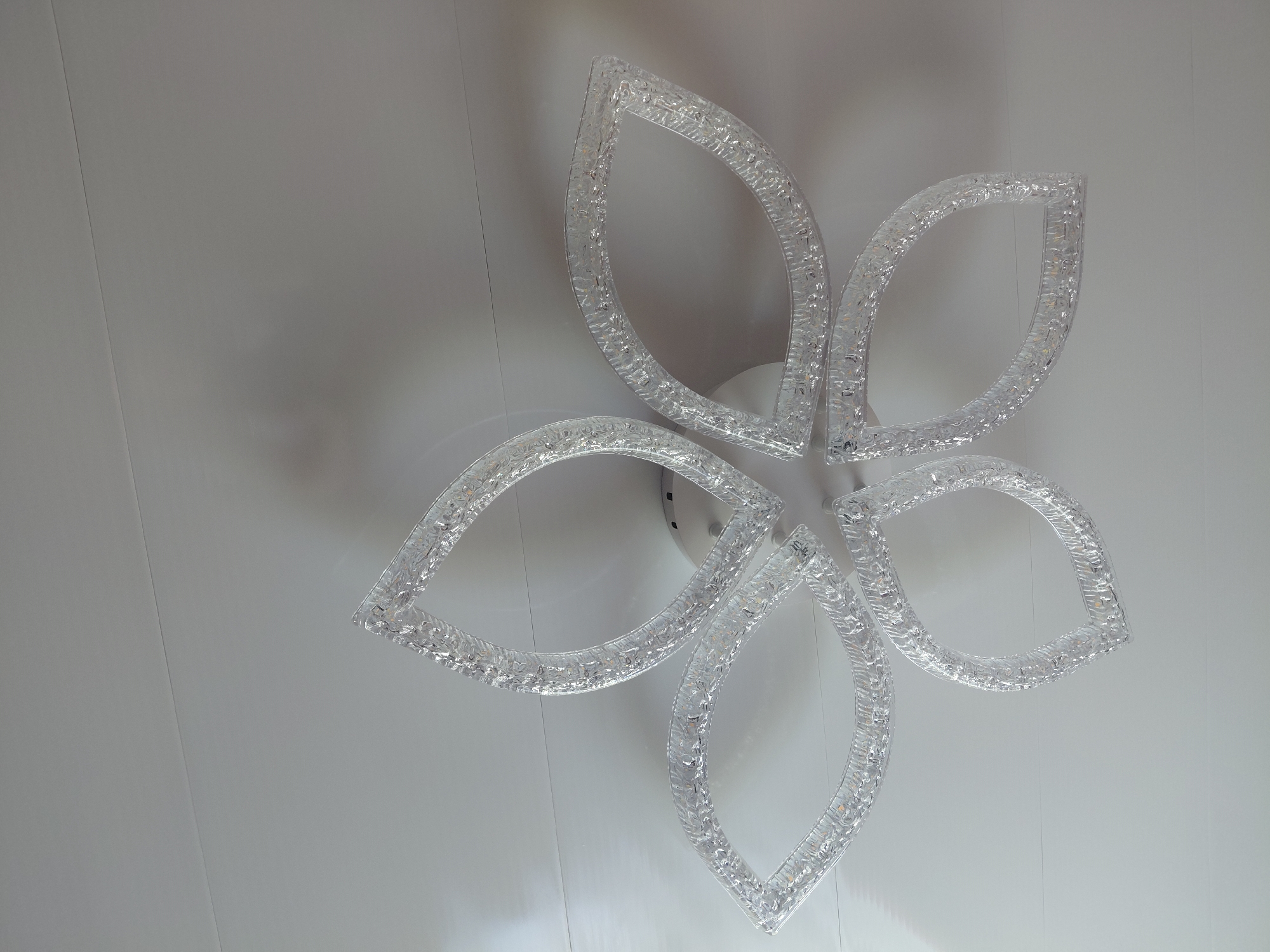 The image shows a close-up, angled view of a modern, flower-shaped ceiling light fixture. The fixture has five petal-like elements, each with a translucent, textured surface that appears to be made of clear acrylic or crystal, embedded with small LED lights. The petals are arranged in a radial pattern around a central, white circular base, which is attached to a plain, white, paneled ceiling. The lighting is even, with soft shadows cast on the ceiling. The overall impression is of a contemporary and elegant interior lighting design. No people or other objects are visible in the image, and the scene is entirely focused on the light fixture and the ceiling. The location in Malinovka, Russia is not visually represented in the image.