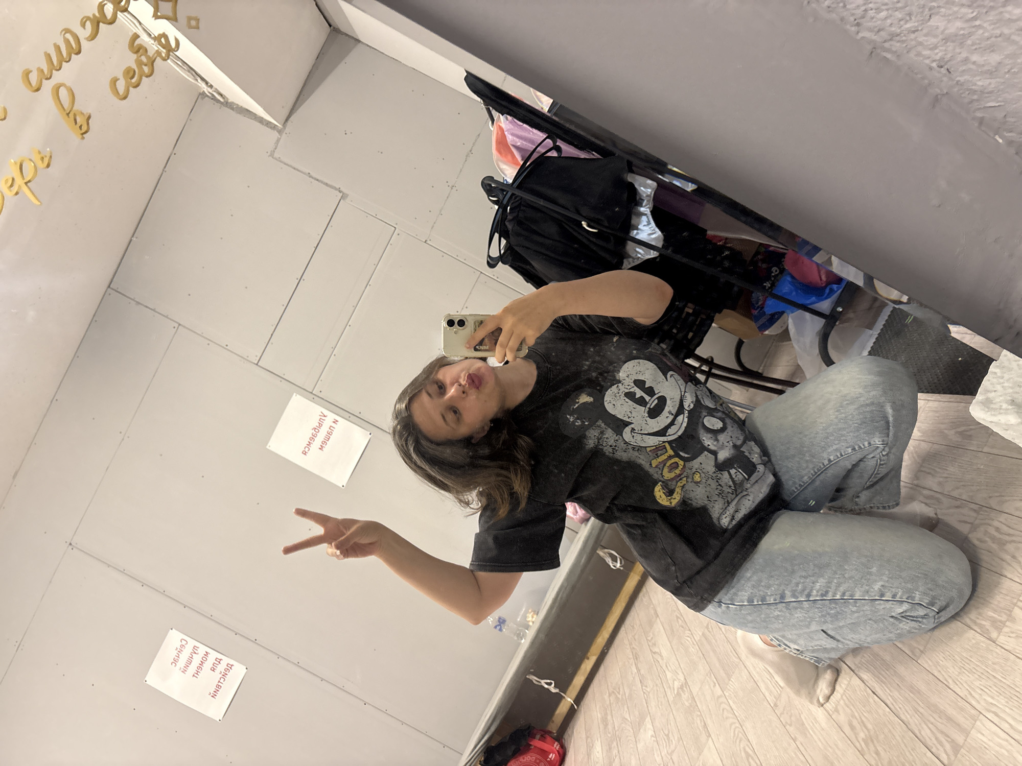 The media file shows a young woman taking a mirror selfie in what appears to be a room with white, panelled walls and a wooden floor. She is wearing a black t-shirt with a graphic of Mickey Mouse and light-wash denim jeans. The woman is kneeling on the floor and making a peace sign with her left hand, while holding her phone in her right hand to take the photo. Her facial expression is playful.
In the background, reflected in the mirror, there are several objects, including a black backpack, some clothing items in various colors, and a metal rack. There are also two white pieces of paper attached to the wall, one on the left and one on the right, with text that appears to be in Russian. The paper on the left has Cyrillic text that reads "СЕЙЧАС", "ПАМЯТИ", "ЧАЙКОВСКОГО", and "ДЛЯ". The paper on the right has Cyrillic text that reads "ПРИВЕТ", "МОМ", and "ЗА". Above her, also reflected in the mirror, there is gold lettering on the wall that reads "в себе" and "сможешь".
The lighting suggests it could be daytime, as the room is well-lit. The overall scene conveys a casual and informal moment. The presence of the text "ЧАЙКОВСКОГО" on the paper provides a strong clue to the location being Chaikovsky, Russia.
