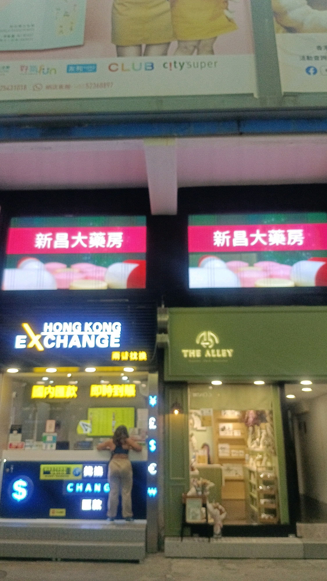 The image captures a street-level view of two adjacent shops at night in Hong Kong, China. The shop on the left is a currency exchange establishment with prominent neon signage. The signage includes the words "HONG KONG EXCHANGE" in English and Chinese characters, along with a large blue dollar sign. Vertical neon signs displaying currency symbols like the Japanese Yen, British Pound, US Dollar, Euro, and Korean Won are visible on the right side of this shop's entrance. Inside, a person is standing behind the counter, facing away from the camera, appearing to be interacting with the exchange services. The shop on the right is a retail store with a green facade and a logo that reads "THE ALLEY" above a stylized symbol. The interior of this shop is visible through the glass, revealing shelves stocked with merchandise and a cashier at the counter. Above both shops, illuminated signs with Chinese characters in red boxes read "新昌大藥房" (Xin Chang Da Yao Fang), which translates to "Xin Chang Pharmacy". In the background, above the pharmacy signs, a larger billboard or banner is visible with English text including "CLUB" and "citysuper", indicating branding for retail or services. The overall environment appears to be a commercial street at dusk or night, with artificial lighting illuminating the shop fronts.