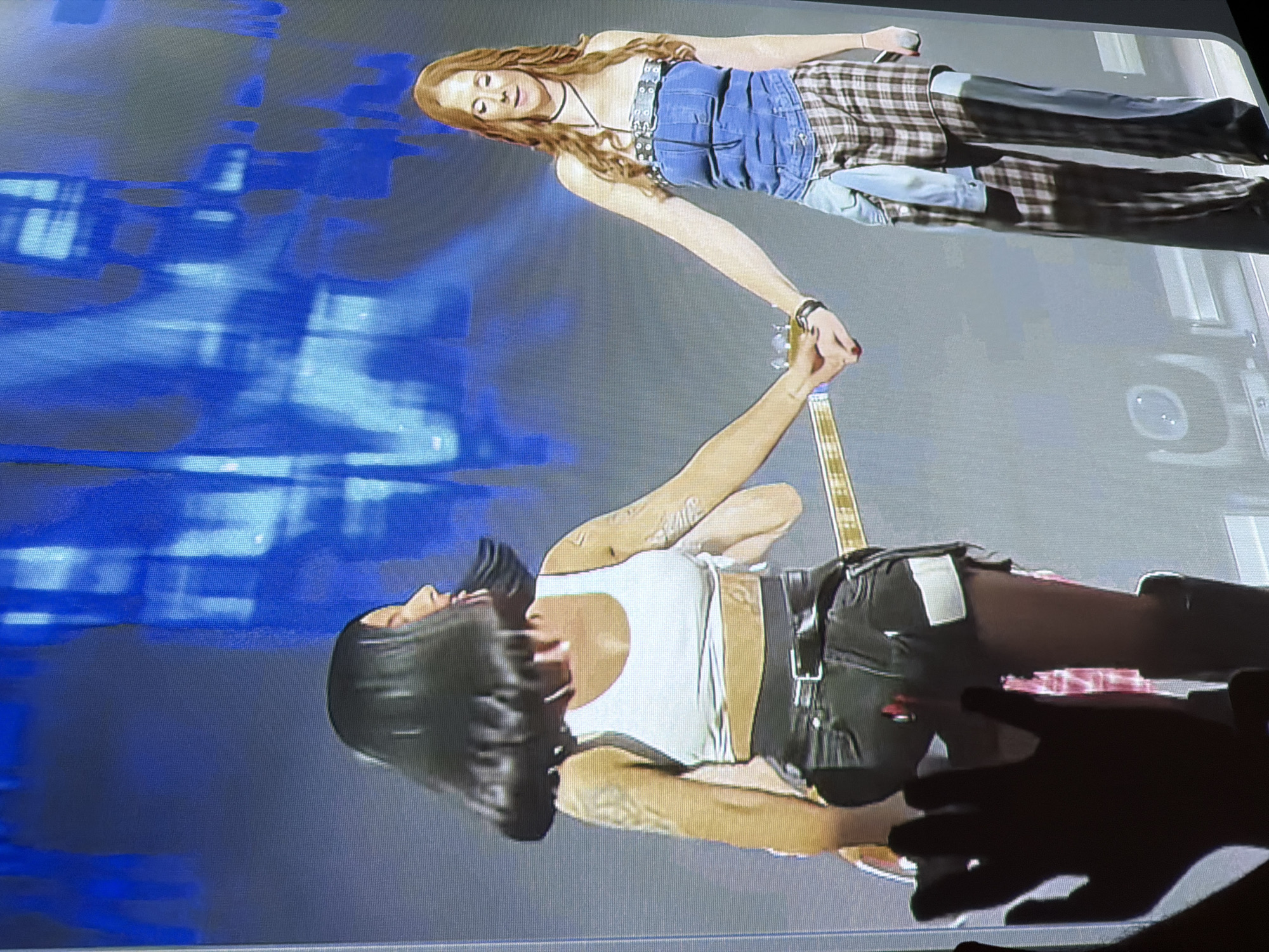Two women are holding hands on a stage with a blue backdrop. The woman on the left has dark hair and is wearing a white crop top and black shorts. The woman on the right has long, red hair and is wearing a denim strapless top and plaid pants. A guitar is visible behind the woman on the left. The scene appears to be a live music performance, possibly indoors. There is a blurred hand in the foreground, suggesting a spectator is taking a photo.