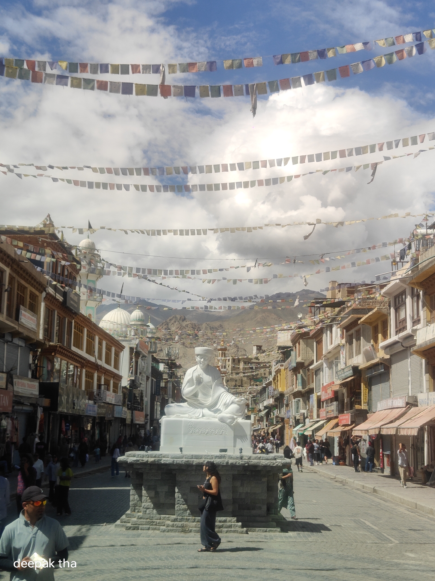 The image captures a street scene in the city of Leh, India, characterized by its unique architecture and cultural elements. In the foreground, a large white statue of a meditating figure is prominently displayed on a stone pedestal. Several people are visible in the vicinity of the statue and along the street, engaging in various activities such as walking, standing, and looking at their phones. The street is lined with multi-story buildings exhibiting a mix of traditional and modern architectural styles, with shops and businesses on the ground floor. Prayer flags, in vibrant colors, are strung across the street and between buildings, creating a festive and spiritual atmosphere, which is characteristic of Leh. In the background, the rugged, mountainous terrain surrounding Leh is visible under a partly cloudy sky. The overall impression is that of a bustling town square, where daily life unfolds against a backdrop of cultural and natural beauty.