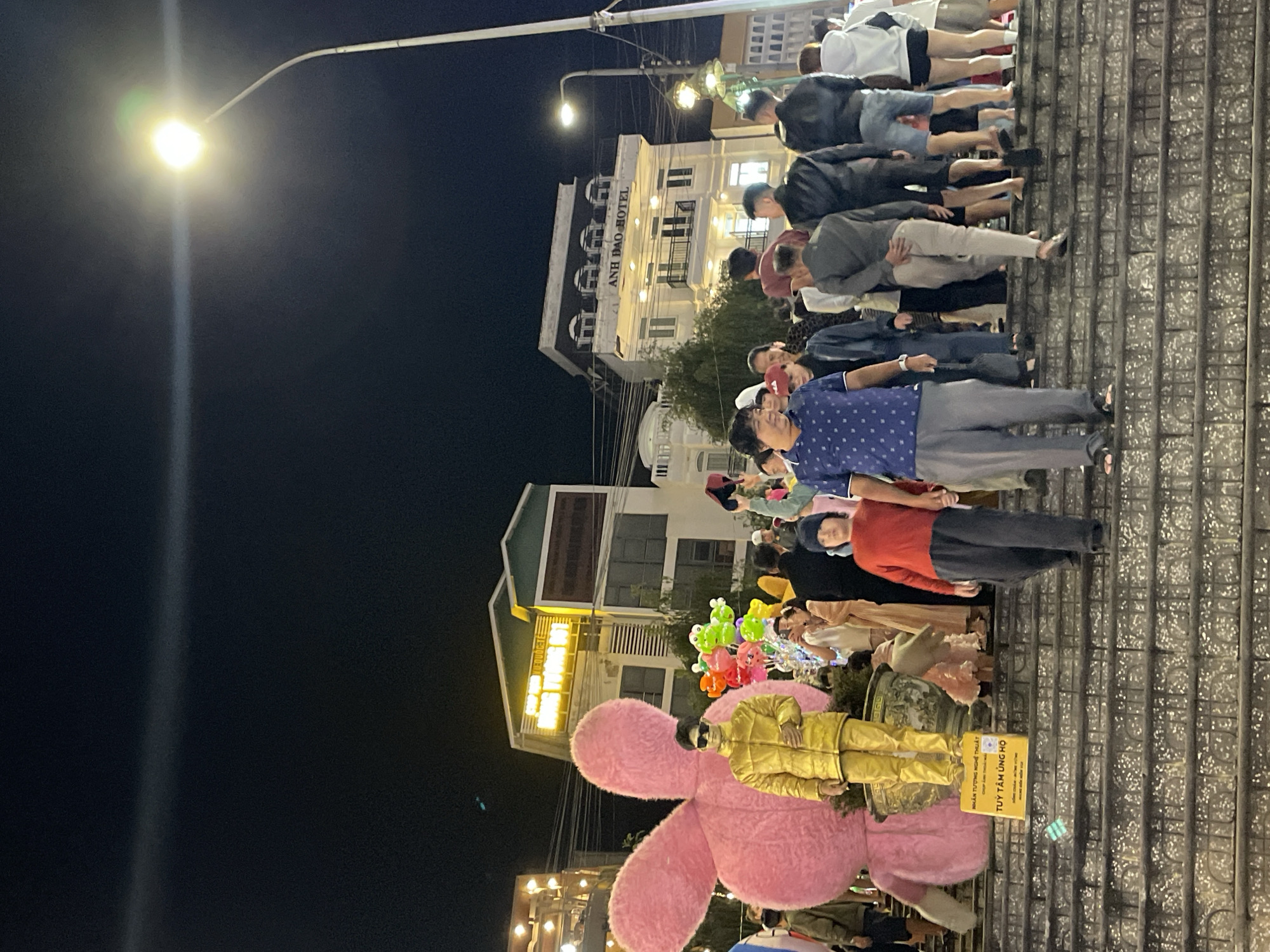 The image captures a nighttime scene on a wide outdoor staircase in Da Lat, Vietnam, likely near a commercial area judging by the buildings visible. The ambiance is festive and crowded, with many people ascending or descending the steps. Dominating the left side is a large, plush pink rabbit statue, serving as a prominent visual anchor. In front of the rabbit, a person dressed in a shiny gold jacket and pants, wearing sunglasses, stands posing, possibly for photos. Behind them, a cluster of colorful balloon animals adds to the celebratory atmosphere.
Several groups of people are scattered throughout the staircase. In the foreground, a man in a blue polka-dot shirt and gray pants walks towards the camera, while a child in a red shirt and dark pants walks down the stairs away from the camera. Other individuals are seen in various states of movement and interaction, some walking, some standing, and some appearing to converse. The background reveals multi-story buildings, one of which is labeled "ANH DAO HOTEL." Streetlights and building lights illuminate the scene, creating a somewhat hazy night atmosphere. The stairs themselves are made of stone and appear well-maintained, with decorative elements incorporated into their design. The overall impression is one of a lively public space bustling with activity at night.