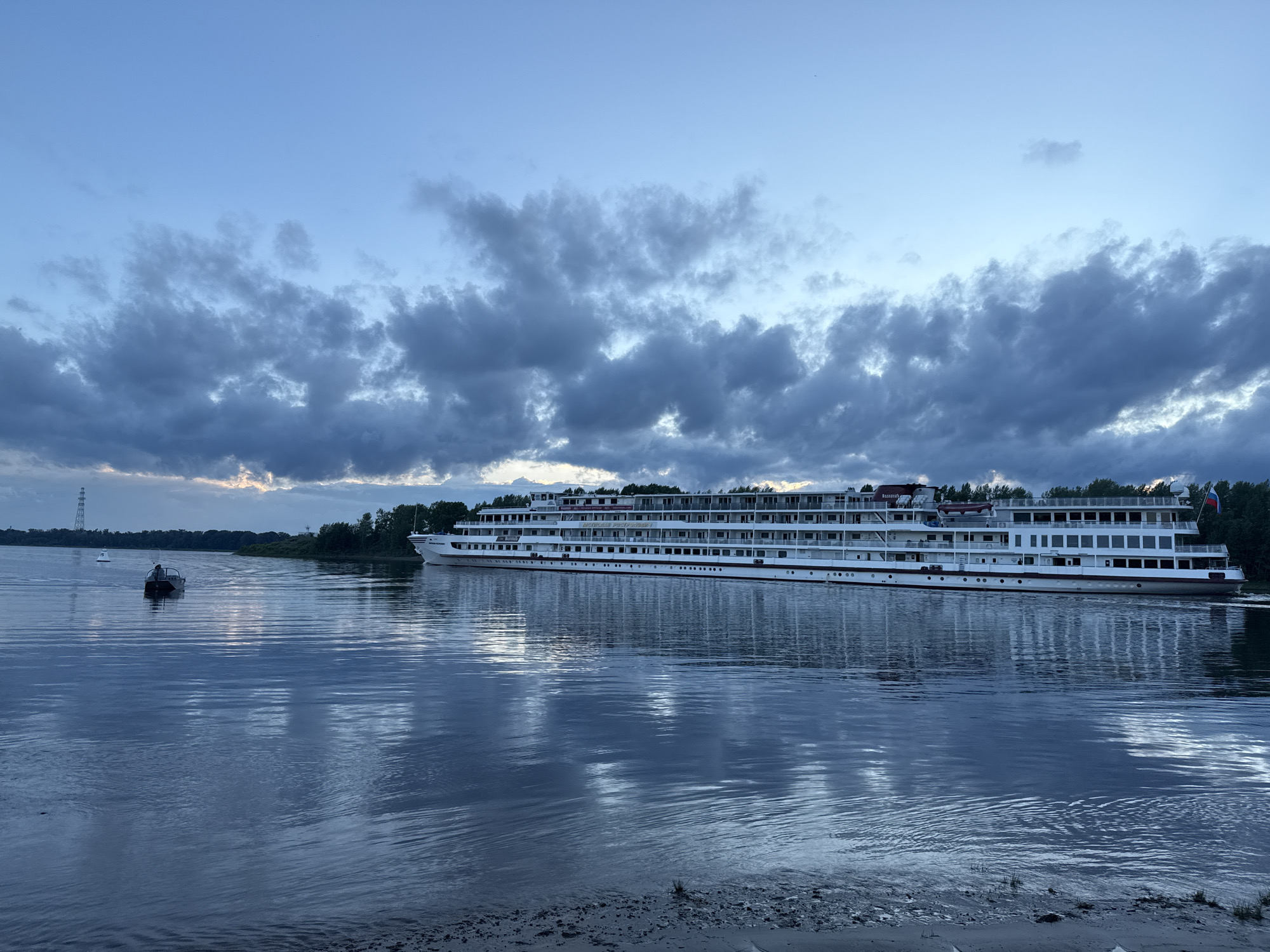 An evening scene shows a large white cruise ship docked on a wide river. The sky is a deep blue with dramatic, dark clouds. The river's surface reflects the ship and the sky, creating a shimmering effect. On the left side of the river, a small boat with one person is visible. The shoreline in the foreground is sandy with some vegetation. Trees line the distant bank.