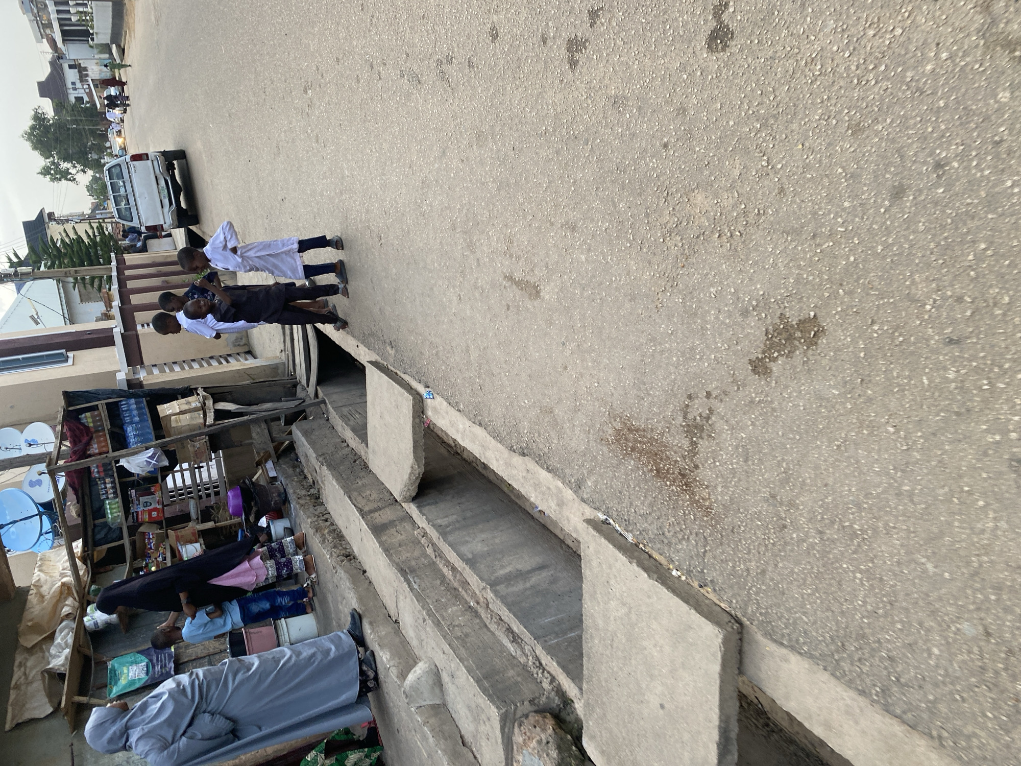 The image captures a street scene in Jos, Nigeria, likely during the daytime. In the foreground, a concrete drain runs along the edge of the asphalt road. To the left, a woman in a grey hijab stands near a makeshift stall with shelves stocked with various items, including bottled drinks and packaged goods. She is interacting with a young boy wearing a light blue shirt and jeans. Another woman in a black hijab is also present at the stall.
Further down the street, a group of four young boys in traditional white attire are standing on the roadside, facing the street. One boy in the foreground is looking towards the camera. A white pickup truck is parked on the right side of the road. In the background, residential buildings and trees are visible, suggesting a typical urban or suburban environment. The overall impression is of a bustling street with everyday life unfolding.