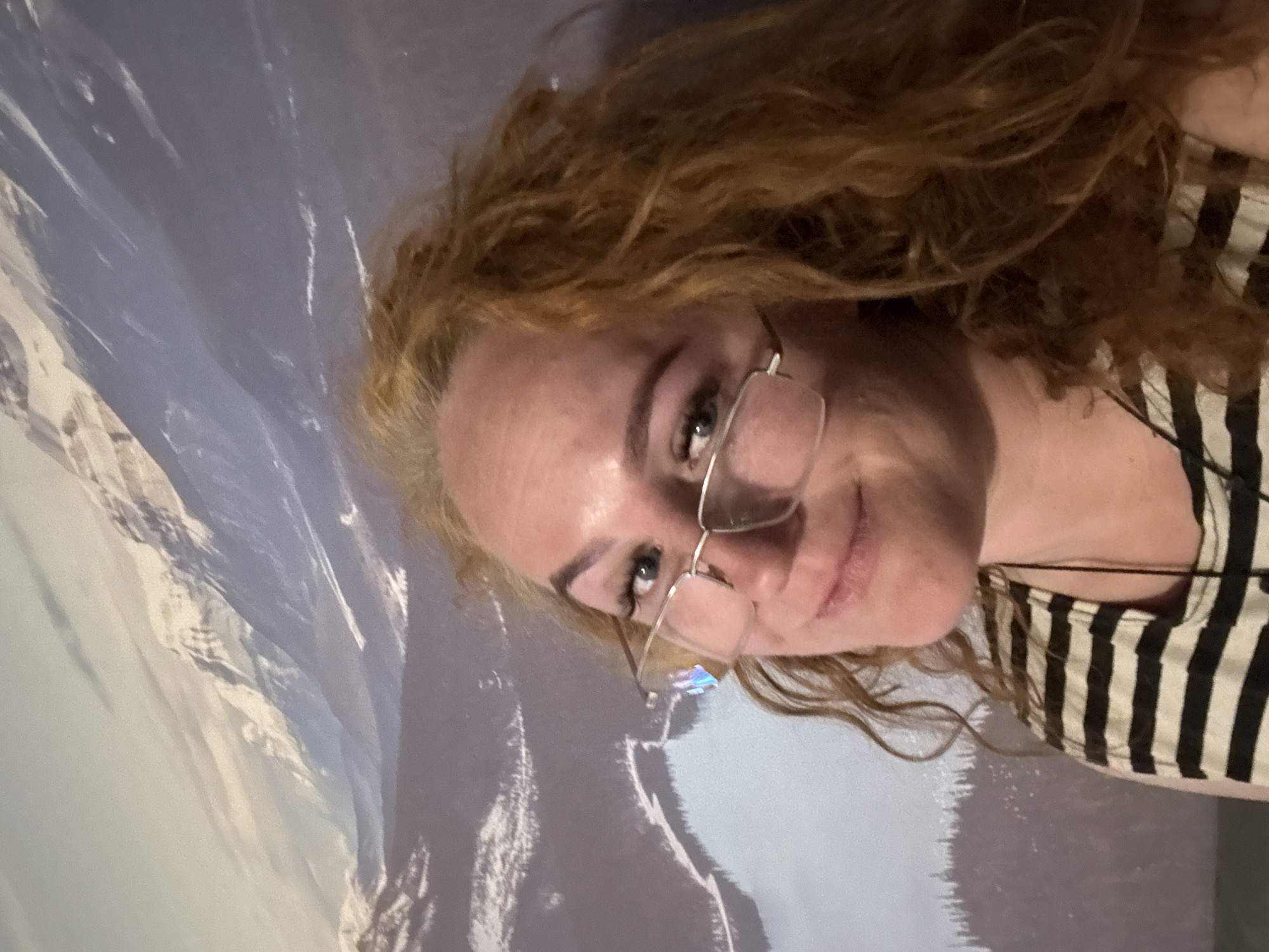 A woman with red hair and glasses is pictured against a backdrop depicting a mountainous scene. She is wearing a striped top. The image appears to be taken indoors, possibly at a location featuring a scenic wall or mural. The location is not discernable as being in Chaikovsky, Russia, or any other particular city based on the visual elements provided.