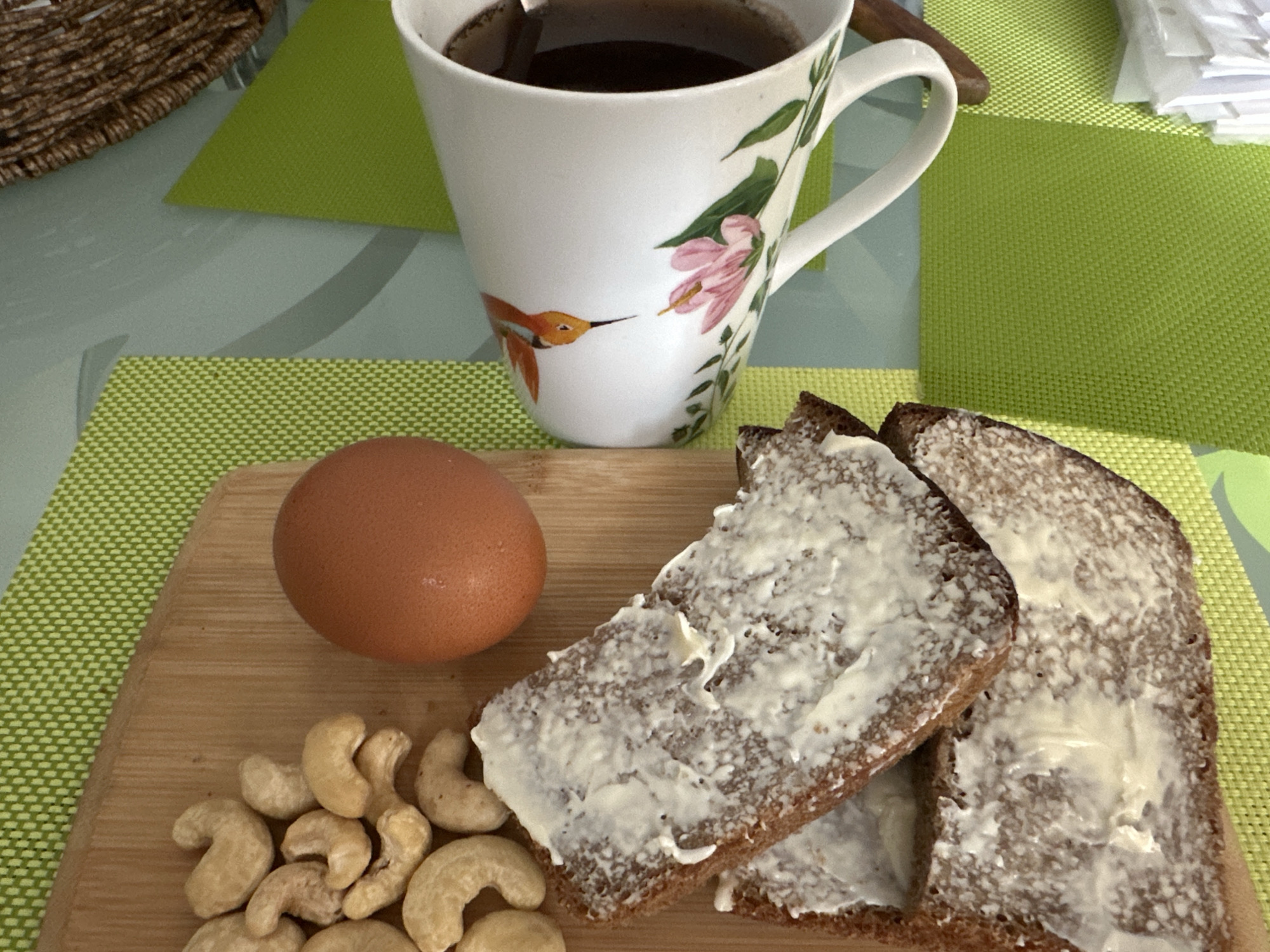 A still life image shows a breakfast spread on a table. In the foreground, a wooden cutting board holds a single brown egg, a pile of cashew nuts, and two slices of dark bread spread with a white substance, possibly butter or cream cheese. Behind the cutting board, a white ceramic mug with a floral and hummingbird design contains dark liquid, presumably coffee or tea. The table is covered with green placemats. A woven basket is visible in the upper left corner. The scene is well-lit, with soft shadows suggesting natural light from a window. The location in Podolsk, Russia is not discernible from the image.