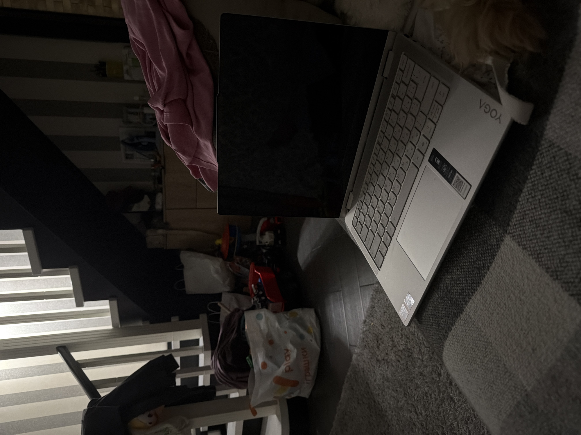 The image depicts an indoor scene, likely a living room or family area, featuring a laptop open on a carpeted floor. The laptop, a silver Lenovo Yoga model, is positioned in the foreground with its screen dark. To the left of the laptop, a white shopping bag with colorful circular graphics and Cyrillic text reading "play игрушки" (play toys) is visible, suggesting the presence of children. Behind the bag, a dark-colored staircase with white railings ascends, and a dark garment hangs on the railing. Further back, shelves hold various items, and a pink blanket is draped over a piece of furniture. The lighting is dim, with vertical stripes of light and shadow on the wall behind the staircase. The floor is a dark wood laminate with a patterned carpet in the foreground. There is no identifiable geographical information suggesting the location is in Bila Tserkva, Ukraine.