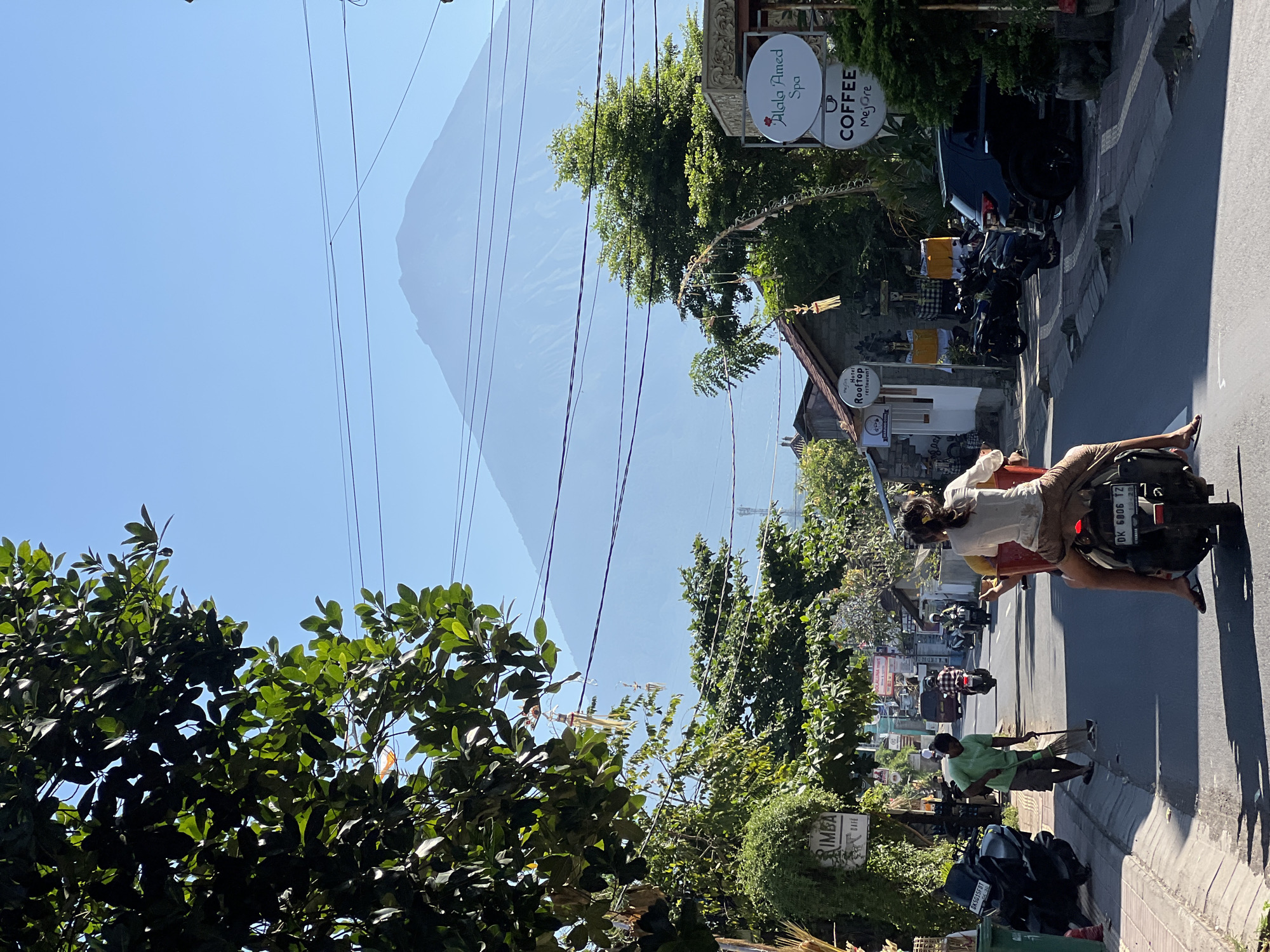 The image captures a street scene in Abang, Indonesia, with a prominent mountain in the background, likely Mount Agung due to the location. The foreground shows a road with several people on motorbikes. Two individuals are riding on a motorbike in the center, with the passenger facing forward. On the left side of the road, a person in a green shirt is sweeping the street. Various shops and signs are visible along the roadside, including "Rimba Cafe", "Hotel Rooftop Restaurant", and "Alala Amed Spa Coffee". Multiple motorbikes are parked on the side of the street, and a black SUV is also present. Trees and overhead power lines are visible throughout the scene, contributing to the urban landscape. The sky is clear and blue.