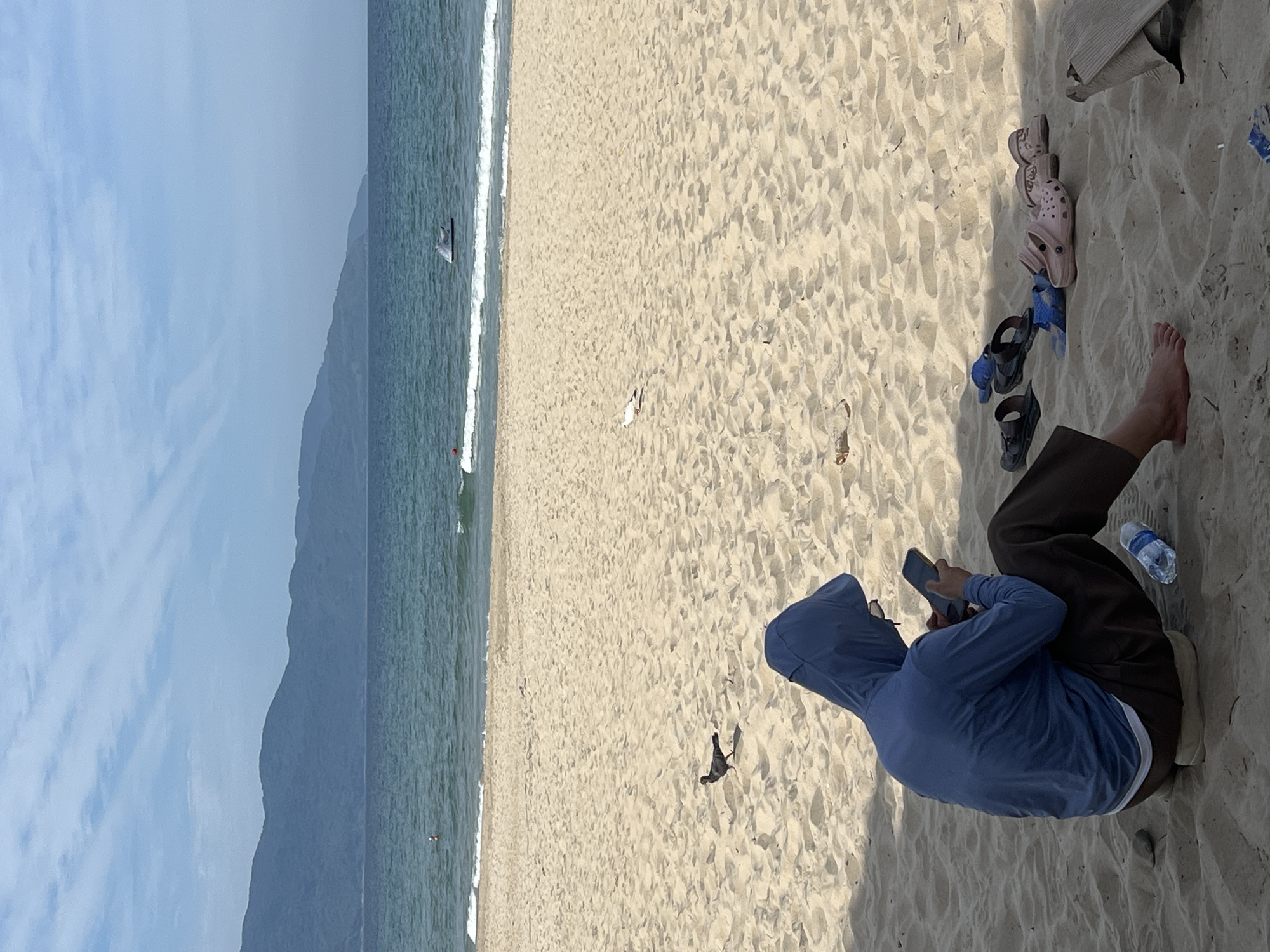 This is an eye-level, medium shot of a person sitting on a sandy beach in Da Nang, Vietnam, looking at their phone. The person is wearing a blue hooded jacket and dark pants, with their feet bare. Several pairs of sandals and flip-flops are scattered on the sand near them, along with a plastic water bottle. A pigeon is visible on the sand to the left of the person, and a seagull is in the background to the right. The ocean stretches out behind the beach, with a gentle surf breaking on the shore. In the distance, mountains line the horizon under a partly cloudy blue sky. A jet ski is visible on the water.