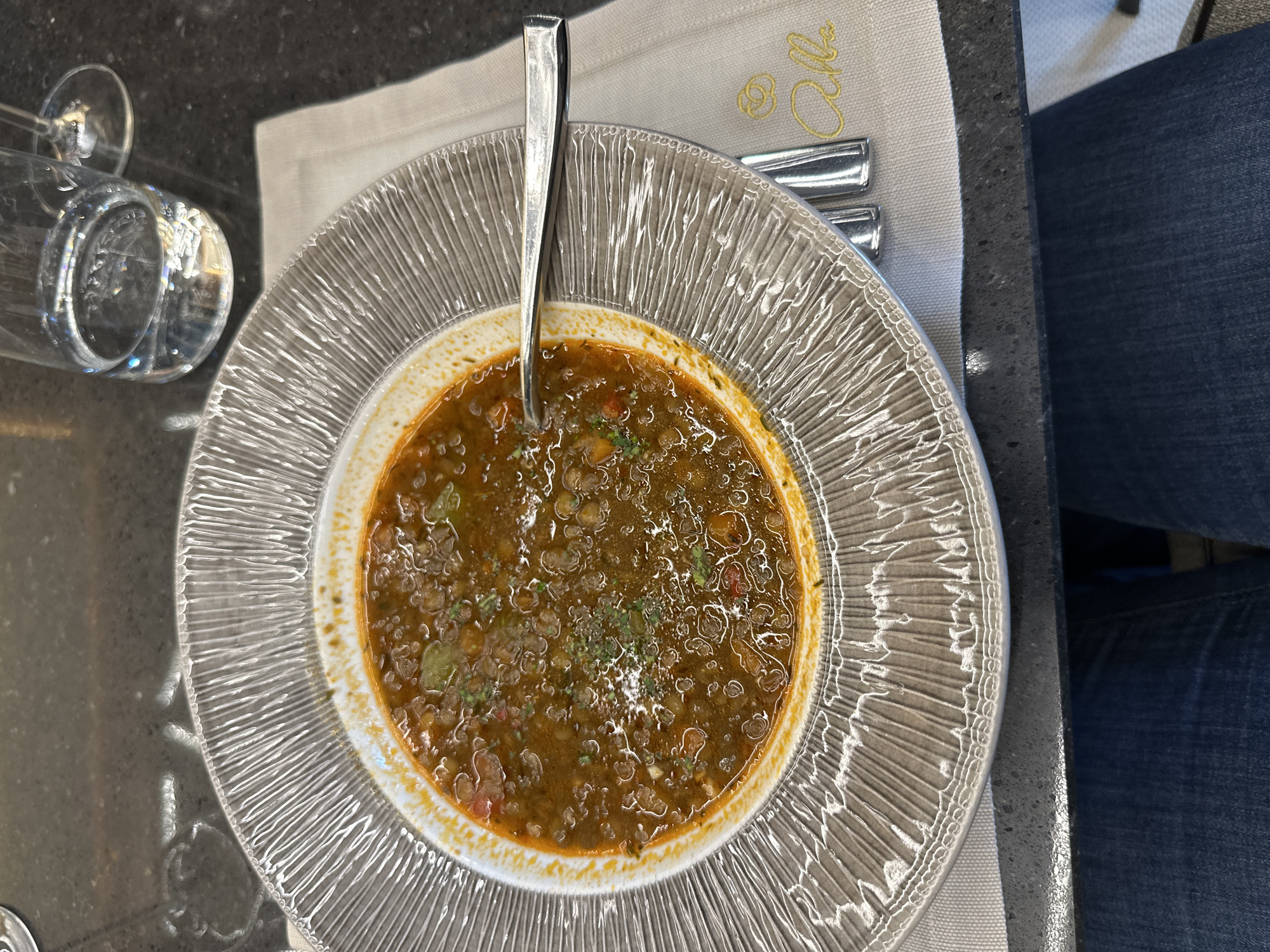 An overhead shot captures a bowl of soup, likely lentil or bean, positioned centrally on a dark, speckled table surface. The soup, a rich reddish-brown, contains visible pieces of vegetables, possibly tomatoes and bell peppers, and is garnished with green herbs. A silver-colored spoon rests within the soup.
The soup is served in a distinctive clear glass bowl with a textured, ridged exterior and a smooth, white inner rim. Beneath the bowl, a light-colored fabric napkin features a yellow-gold embroidered logo or text that appears to read "Alla" in a cursive script. To the right of the soup bowl, additional silver cutlery is visible on the napkin.
On the left side of the table, a clear wine glass and a tumbler or water glass are present, reflecting ambient light. The bottom right corner of the frame shows a person's leg clad in blue denim jeans, indicating someone is seated at the table. The lighting is bright and natural, suggesting it is daytime. The scene depicts a meal being served in a dining setting, likely indoors at a restaurant or cafe in Kazanlak, Bulgaria.