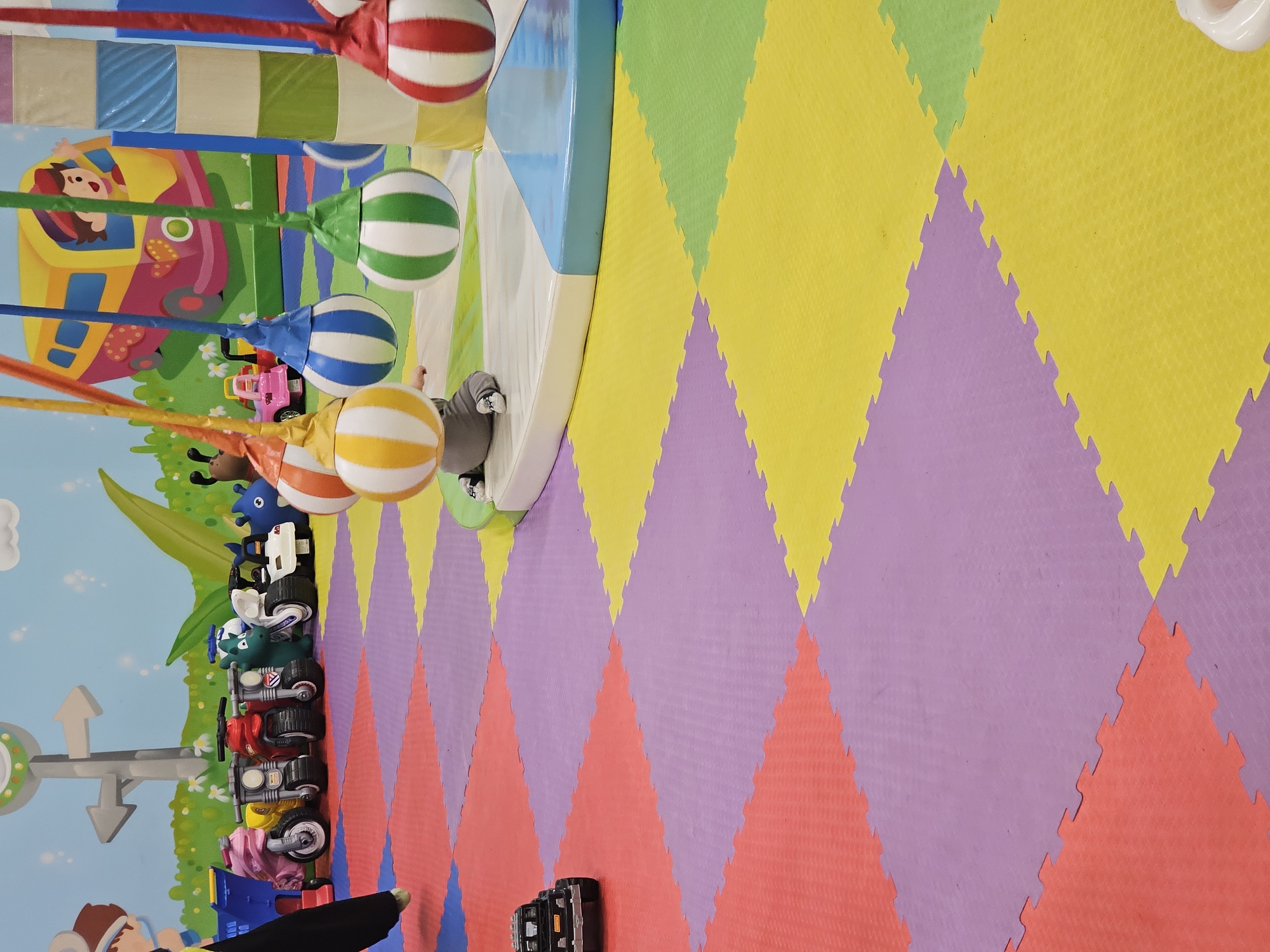 The image depicts an indoor children's play area in Khimki, Russia, characterized by colorful diamond-patterned interlocking foam floor tiles. The tiles are primarily in shades of yellow, green, purple, and red. Adjacent to the tiled floor is a white and blue platform with striped balls suspended from above, likely part of a play structure. A child's legs are visible beneath the platform. On the wall, there are cartoonish decorations including vehicles and characters. There is also a row of toy motorcycles lined up. At the bottom, a person's hand and an electronic device are visible.
