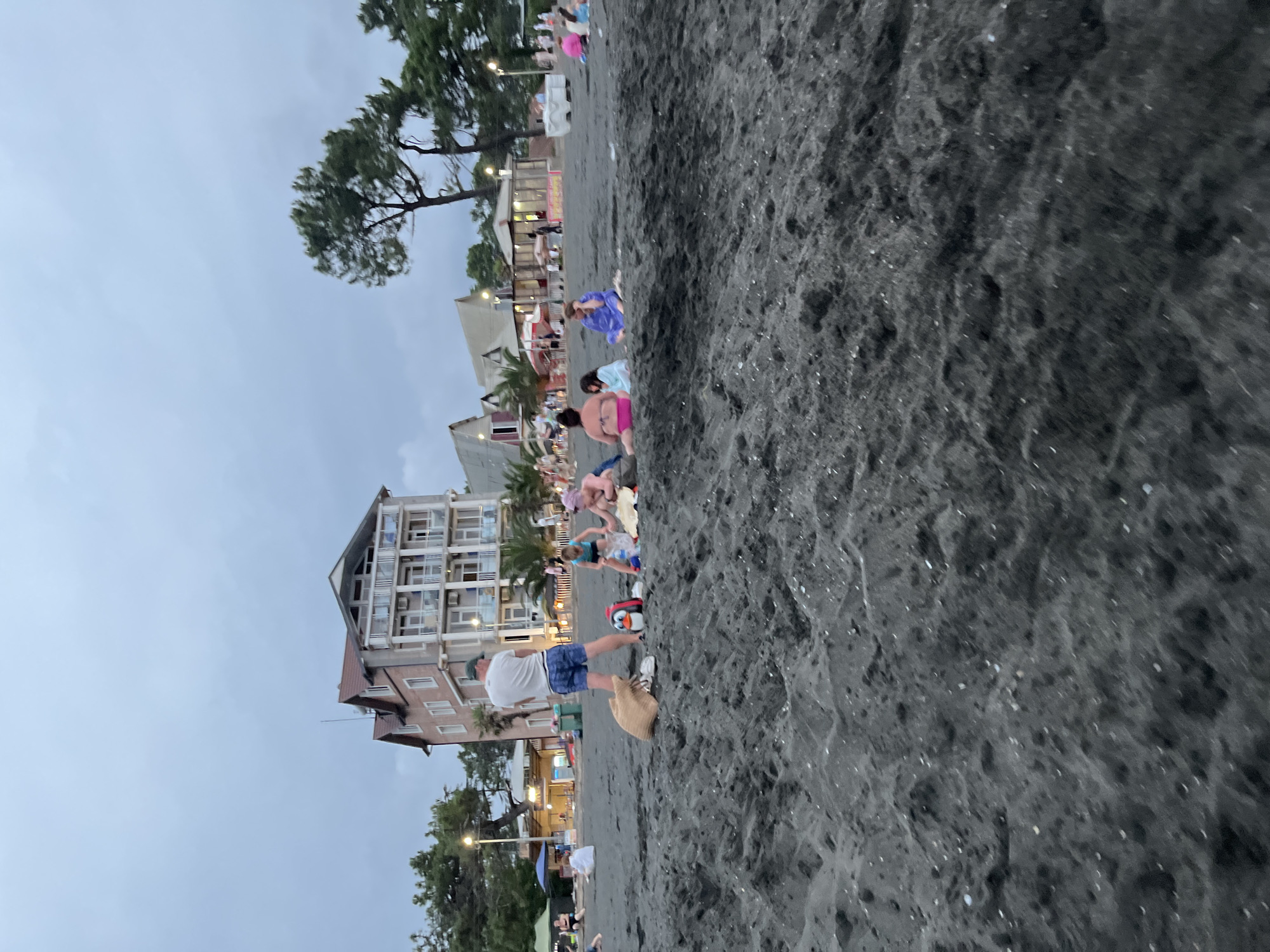 The image shows a crowded beach scene in Ureki, Georgia. The beach has dark, sandy shores, and several people are gathered in small groups on the sand. In the background, there is a multi-story building with balconies, characteristic of a hotel or resort in the area. Trees and other structures, possibly shops or cafes, line the edge of the beach. The sky is overcast, suggesting it might be late afternoon or early evening. Some people are sitting on blankets, while others appear to be standing and interacting. There are beach bags and a distinctive penguin-shaped toy visible on the sand. The overall atmosphere suggests a leisurely day at the beach.