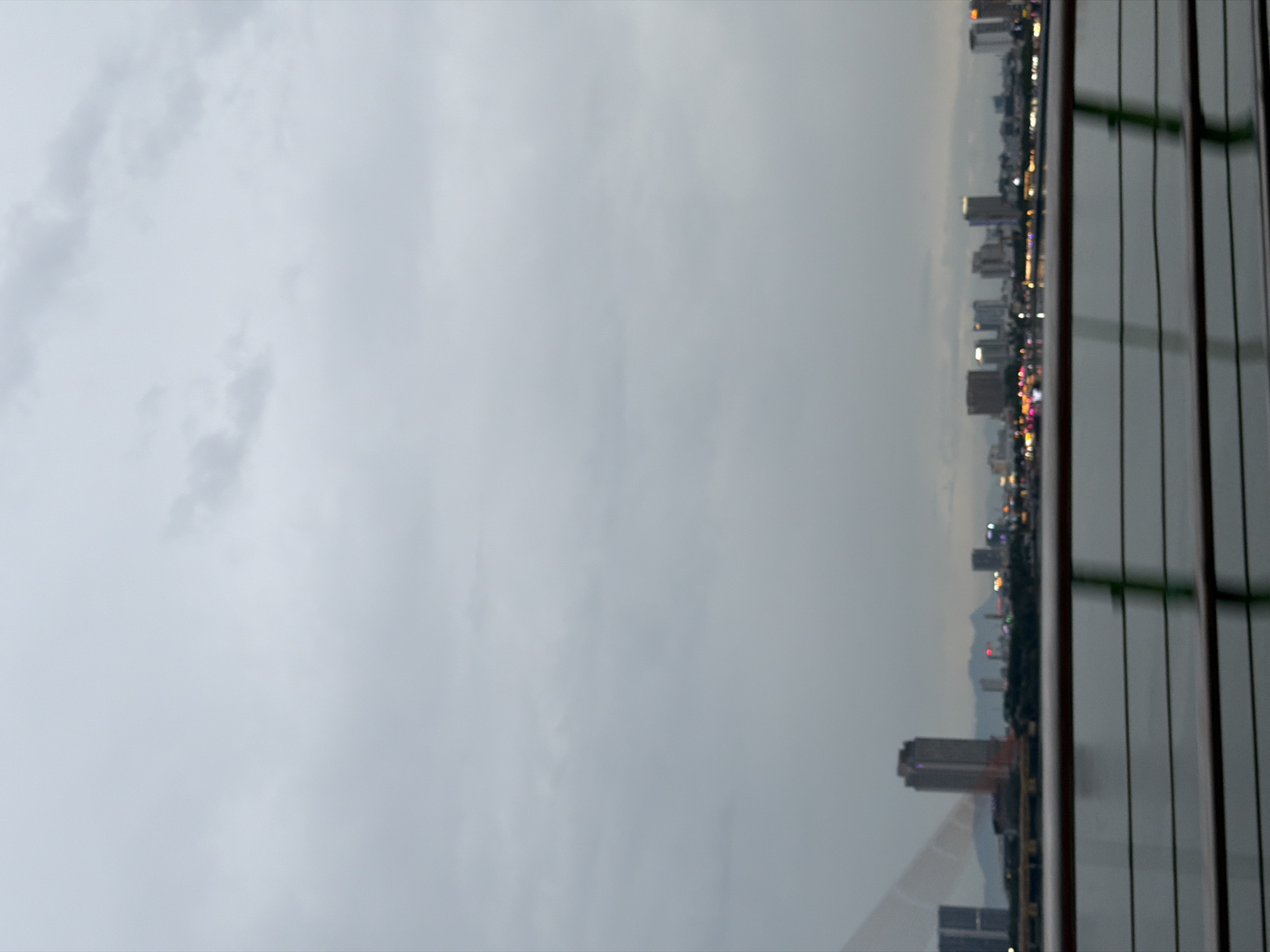 The image shows a blurry, overexposed daytime view of the Đà Nẵng skyline across a body of water. In the foreground, horizontal railings, likely from a bridge or elevated walkway, are visible. The skyline features several tall buildings, including a prominent skyscraper with a pointed top, as well as lower structures and a hint of distant mountains. The sky is overcast and grey. The water in the foreground reflects some of the city lights, indicating it might be dusk or dawn. The overall impression is of a city waterfront viewed from a moving perspective.