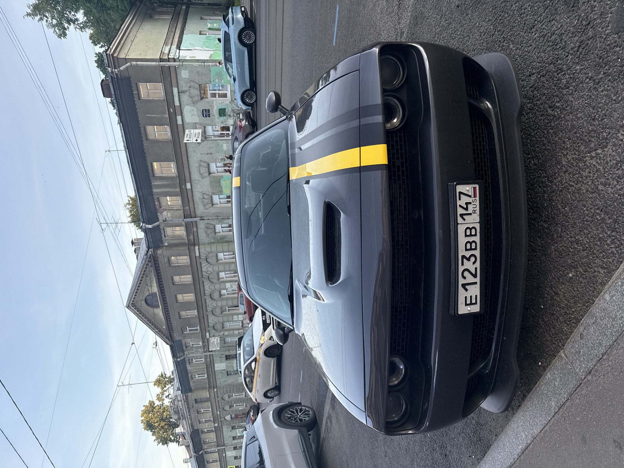 A dark grey Dodge Challenger with yellow racing stripes on the hood is parked on a city street. In the background, there are several older buildings with ornate architectural details. A light blue SUV is parked further down the street, and other cars are visible. The sky is clear and blue, indicating daylight. A Russian license plate, "E123BB 147 RUS", is visible on the front of the Dodge Challenger. The scene appears to be in Saint Petersburg, Russia, given the style of the buildings and the license plate format.