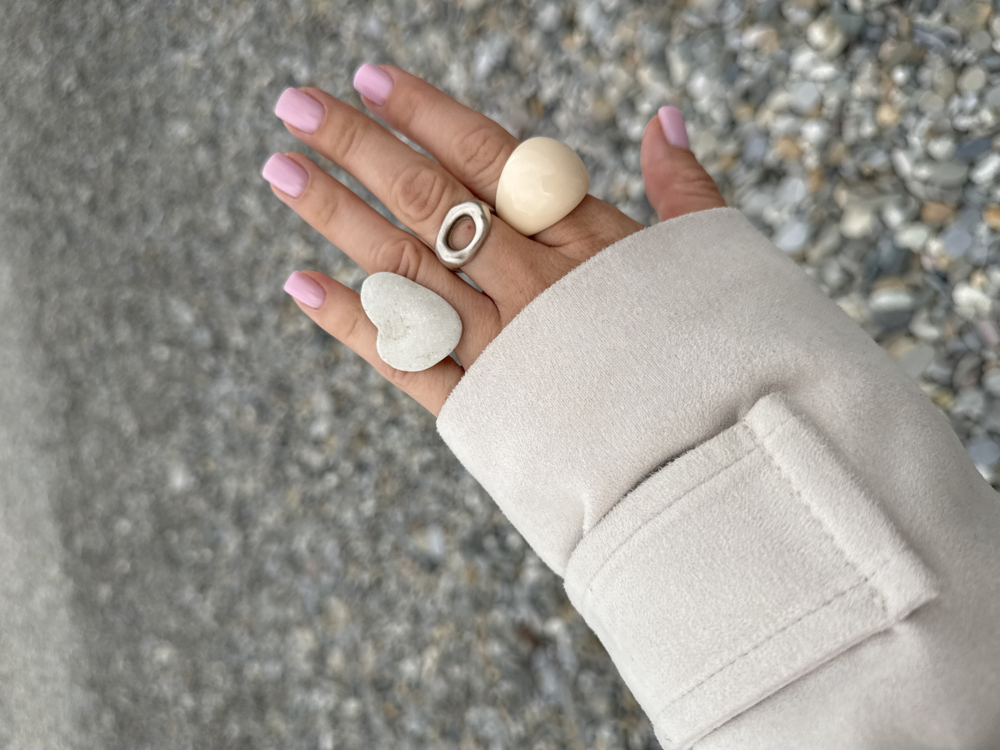 The image shows a person's hand adorned with three rings – one silver with an oval opening, one round and off-white, and one shaped like a heart. The hand is wearing a light beige jacket and is set against a blurry backdrop of gray and white pebbles. The person's nails are painted a light pink color. It is impossible to confirm the specific location in Sochi, Russia, without more information.