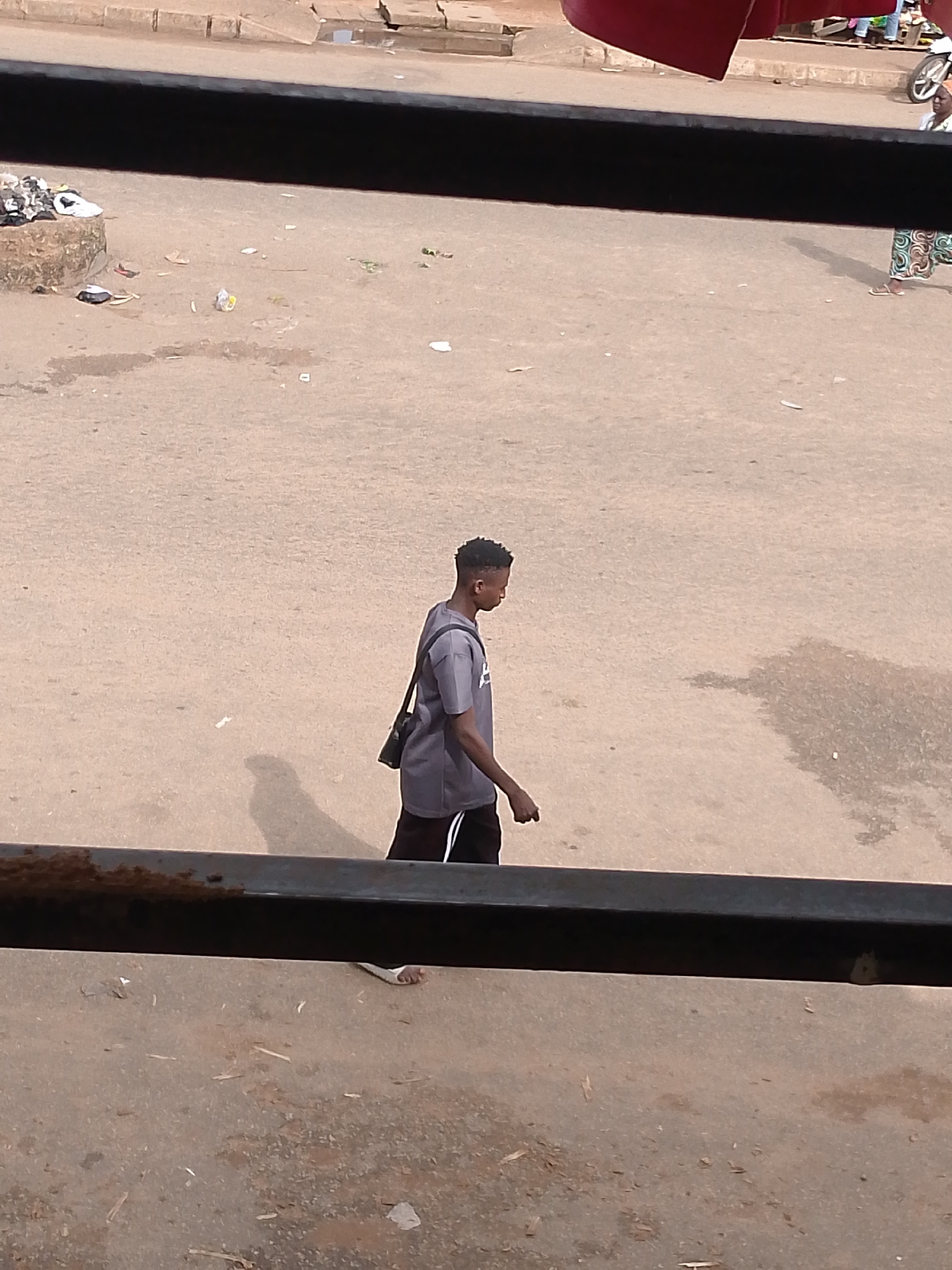 The image shows a young man walking on a dusty, unpaved road in Bukuru, Nigeria. He is wearing a grey t-shirt and dark shorts with white stripes. A dark bag is slung across his shoulder. The road is littered with various pieces of trash, including plastic wrappers and debris. In the background, a curb and a rough concrete structure are visible. The scene suggests an outdoor environment, possibly a market area or a residential street with poor waste management. The overall impression is one of a typical day in a busy, potentially lower-income urban setting.