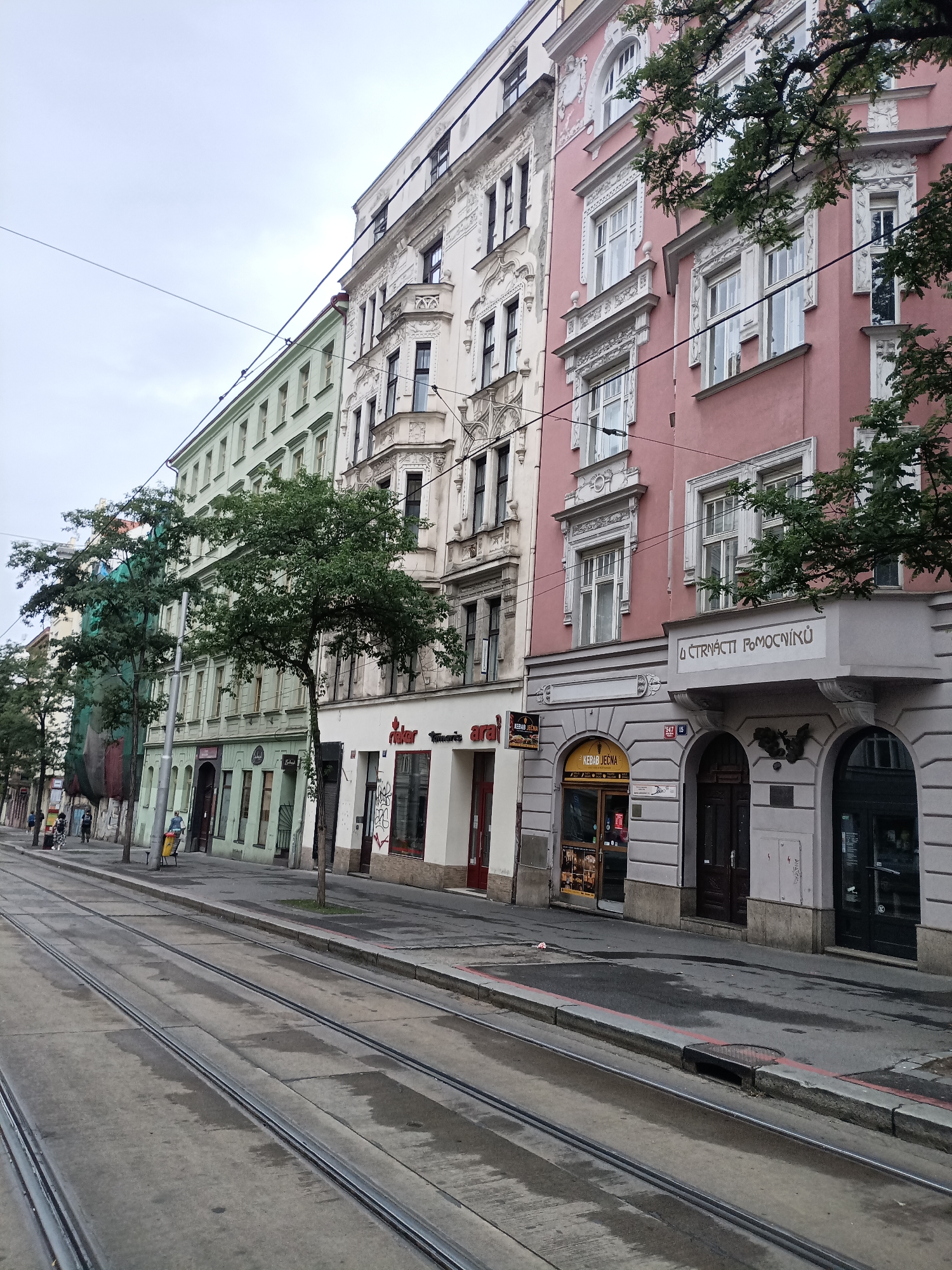 The image captures a street scene in Prague, Czechia, focusing on a row of multi-story buildings lining a tram track. The buildings exhibit distinct architectural styles, with the leftmost structure painted in a light green hue, followed by a white building with ornate detailing, and a prominent pink building further to the right. The facades feature numerous windows, some with decorative moldings and bay windows. Trees are planted along the sidewalk, with one tree on the right side partially obscuring the pink building.
The foreground is dominated by a set of tram tracks embedded in the concrete street, which appears wet in patches, suggesting recent rain. A sidewalk runs alongside the tracks, with various storefronts visible at street level. Signs for businesses such as "Rieser" and "Kebab Ječna" are discernible. A few individuals are present on the sidewalk, including one person pushing a yellow cart and another walking with a companion. Overhead, electrical wires for the trams crisscross the sky, which is overcast. The overall impression is of an urban environment on a cloudy day.