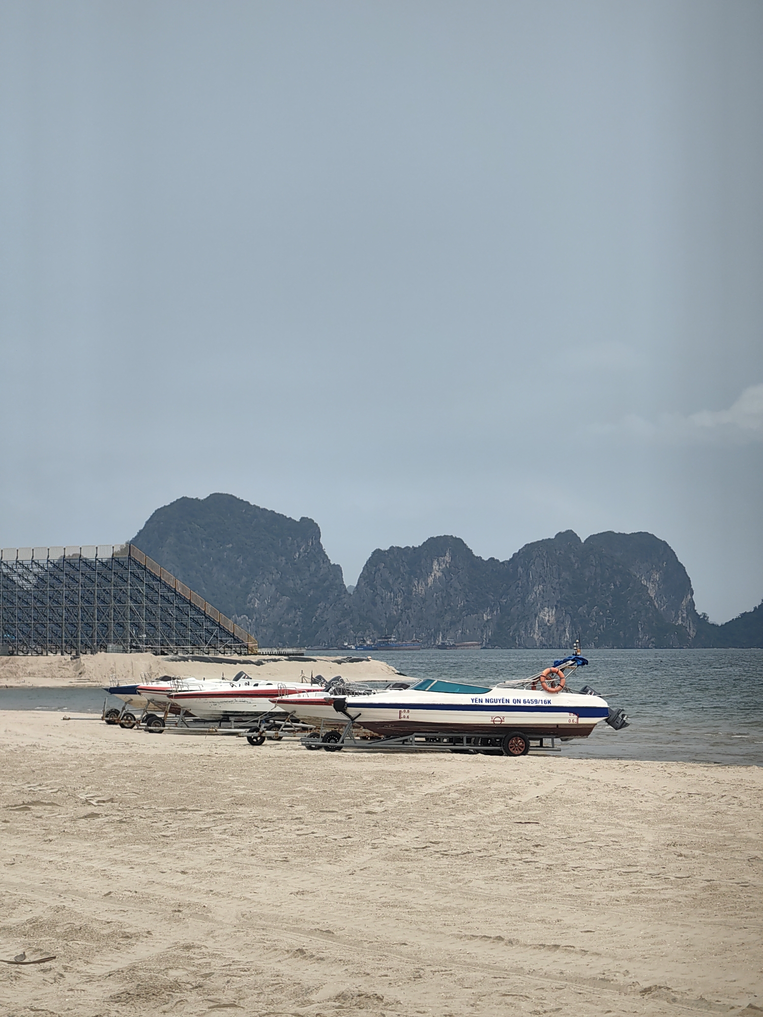 The image shows two motorboats on trailers, sitting on a sandy beach in Bai Chay Ward, Vietnam. In the background, there's an artificial structure made of scaffolding, plus several limestone karst hills, characteristic of the landscape. The sea is visible behind the boats and in front of the hills. The sky is overcast.