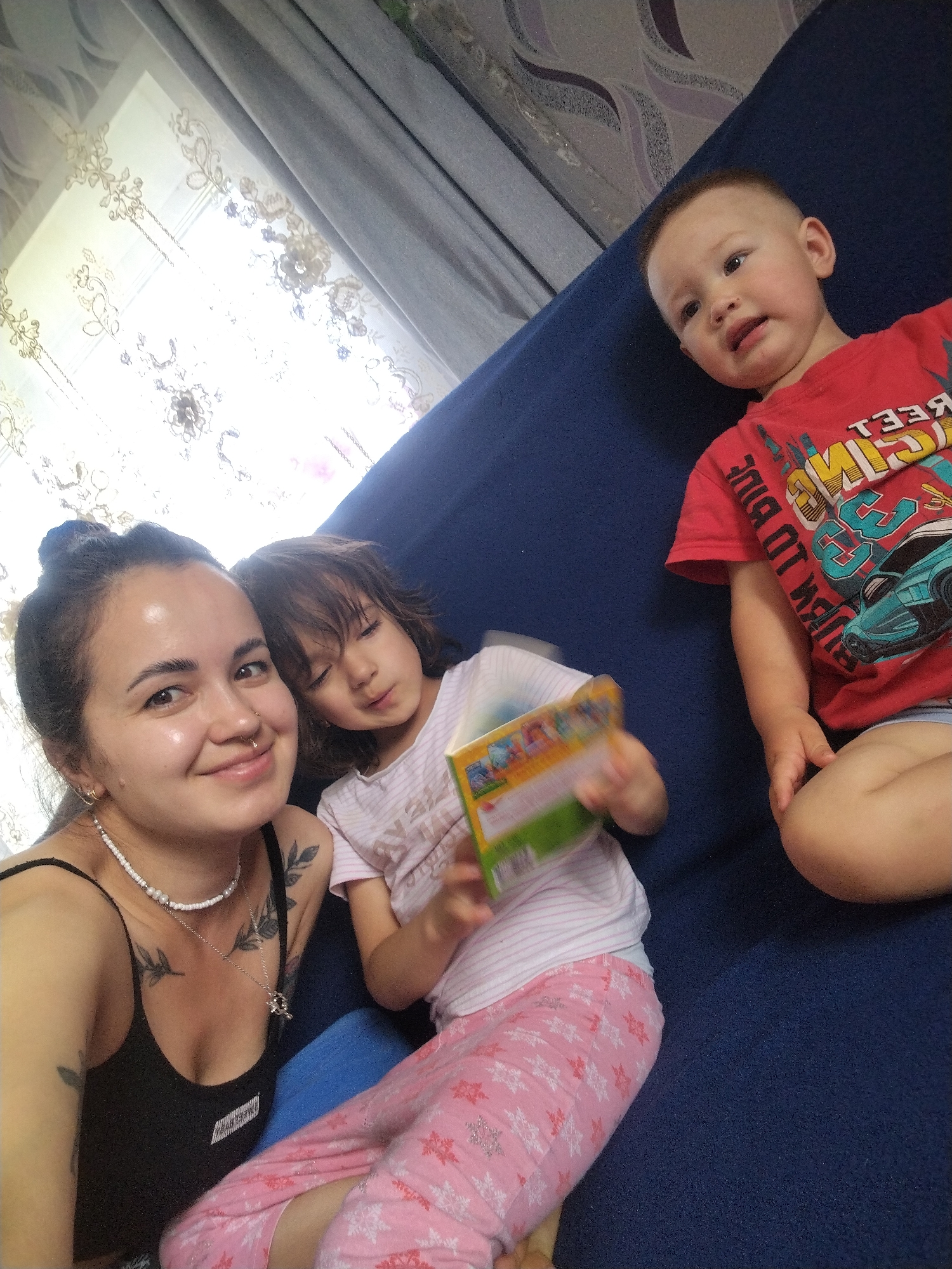 In Гожан, Russia, three people are pictured indoors. A woman with her hair in a bun, wearing a black tank top and a white beaded necklace, smiles at the camera. To her right, a child with brown hair, wearing a pink and white striped shirt and pink snowflake patterned pants, is looking down at a book. To the right of the child, a toddler with short hair, wearing a red t-shirt, looks up and to the left. The three are sitting on a blue sofa. Behind them, a window with sheer patterned curtains is visible, and to the left, a gray curtain is drawn. The overall setting appears to be a domestic environment, possibly a living room.