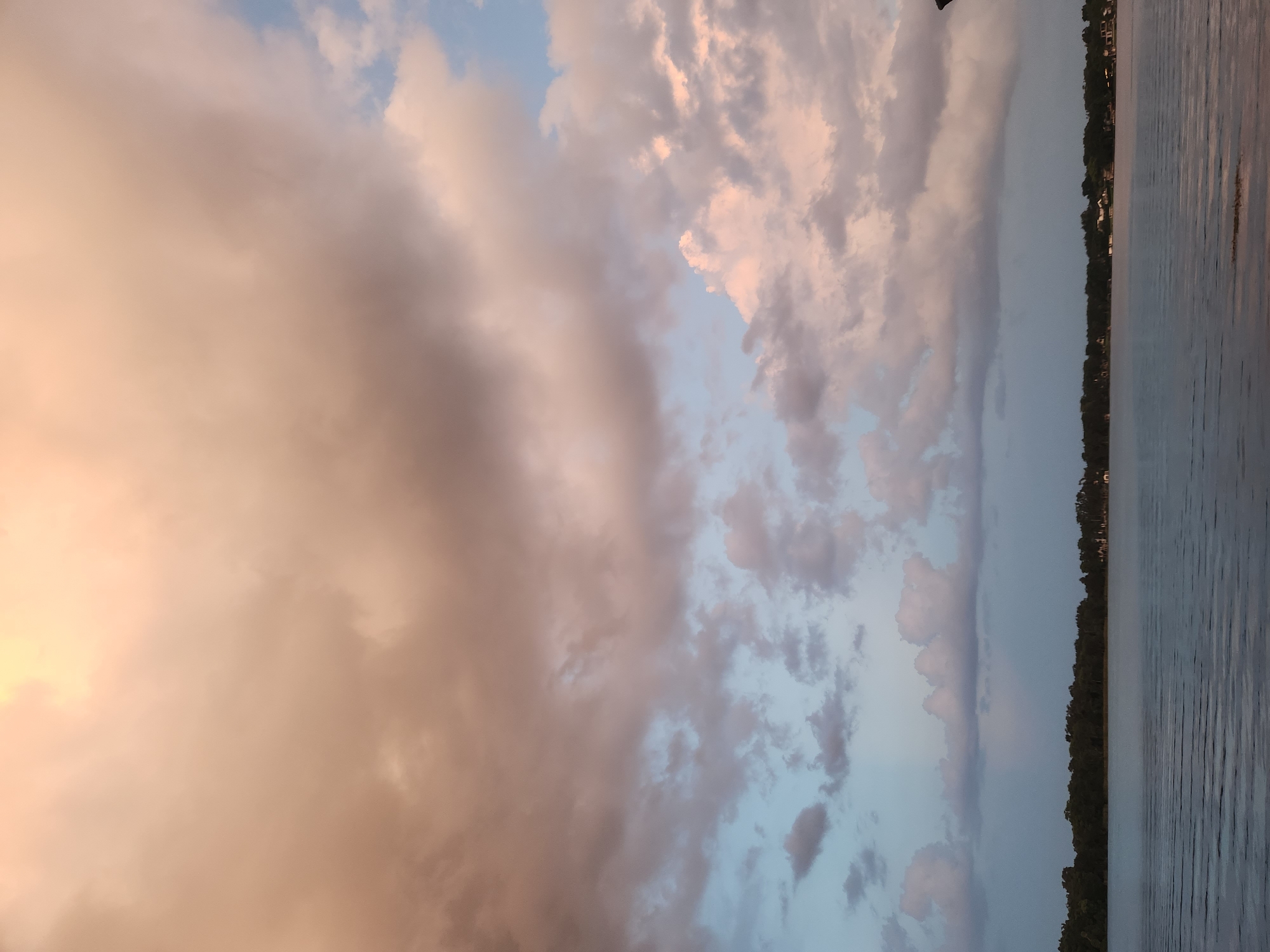 An image captures a wide expanse of the sky, dominated by voluminous clouds tinged with hues of peach, pink, and white, set against patches of pale blue. Below the sky, a calm body of water reflects the colors of the clouds, with subtle ripples disturbing its surface. Along the far bank of the water, a dense line of dark green trees forms a horizon, punctuated by a few scattered white structures that suggest the presence of buildings. The lighting indicates either dawn or dusk, casting a soft, diffused glow over the scene. This view is consistent with locations around lakes in Winter Park, Florida, given the typical landscape and atmospheric conditions common to the area. There are no people or animals visible in the frame.