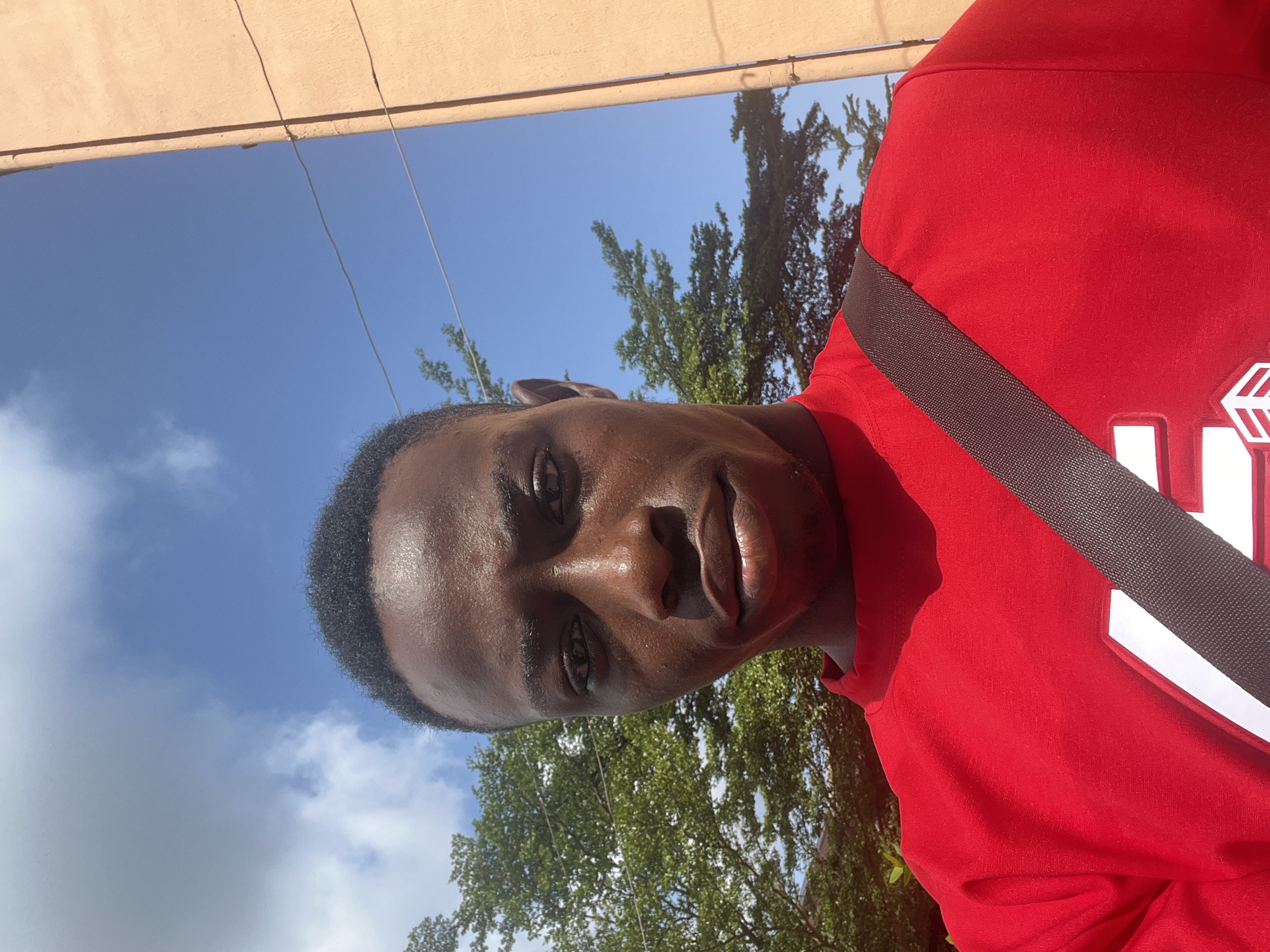 A frontal, eye-level, medium shot captures a dark-skinned Black male in a bright red t-shirt, holding a dark brown strap across his chest. The male's head is tilted slightly down, with his gaze directed at the camera, his expression neutral. His short, dark, curly hair is visible at the top of the frame. The background consists of a bright blue sky with scattered white clouds, a large green tree to the left, and a pale orange building to the right. Electrical wires are visible against the sky, suggesting an outdoor urban environment. The lower portion of the t-shirt features a white logo with red accents. The overall lighting is bright, indicating daytime. The image appears to be a selfie taken outdoors in Nnewi, Nigeria, as indicated by the context provided.