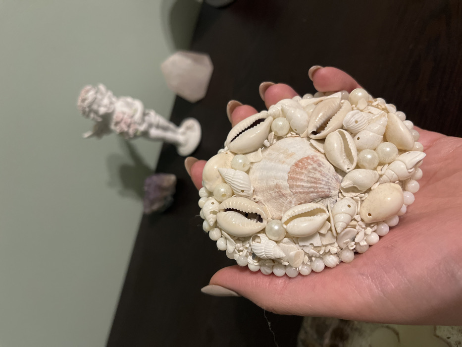 A light-skinned human hand, exhibiting manicured fingernails with light polish, firmly grasps a circular, domed decorative object in the city of Varna, Bulgaria. The object, approximately 10-15 cm in diameter, is densely encrusted with numerous small white, off-white, and beige seashells of varied shapes, interspersed with white spherical beads resembling pearls. A prominent, fluted bivalve shell, exhibiting a subtle pink hue, is situated centrally. The background consists of a dark, horizontal surface on which three items are partially visible: a blurred purplish crystalline rock to the left, a blurred white statuette of a cherubic figure holding an unidentifiable item on a round base in the center, and a blurred light-colored, translucent crystal or mineral formation to the right. The wall behind the surface is a plain, muted green color.