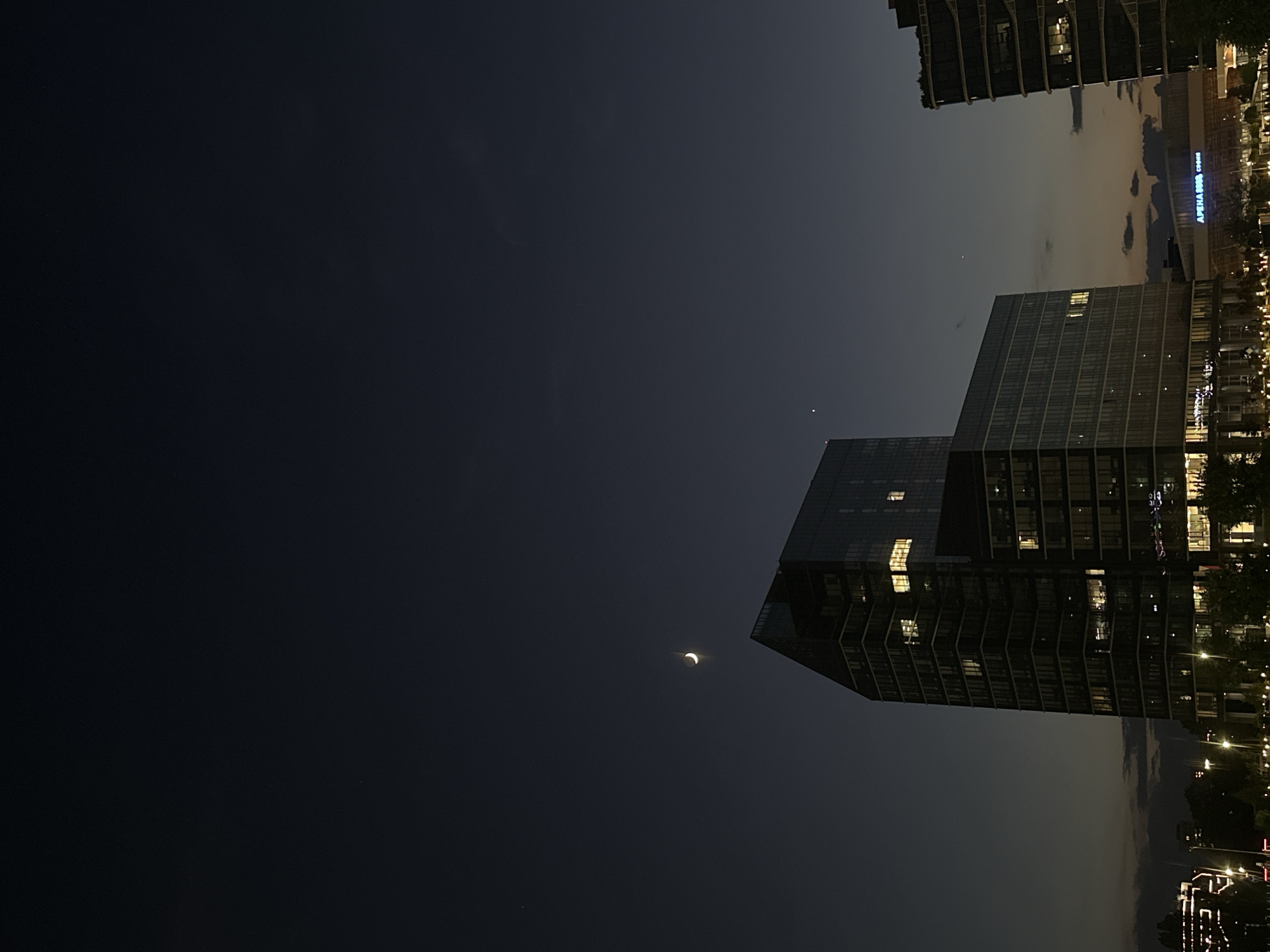 This is a nighttime photograph taken in Sofia, Bulgaria, featuring a crescent moon and a bright star or planet in the dark sky above modern glass-facade buildings. The main building in the foreground is a tall, angular structure with illuminated windows, suggesting occupancy. To the right, another contemporary building with a stepped facade is visible, also showing interior lights. The lower portion of the image depicts a street scene with lampposts, trees, and other structures, hinting at an urban environment. The sky is deep blue, transitioning to a lighter tone near the horizon, with a few scattered clouds. The overall impression is one of a city at dusk or early night, with a clear celestial view.